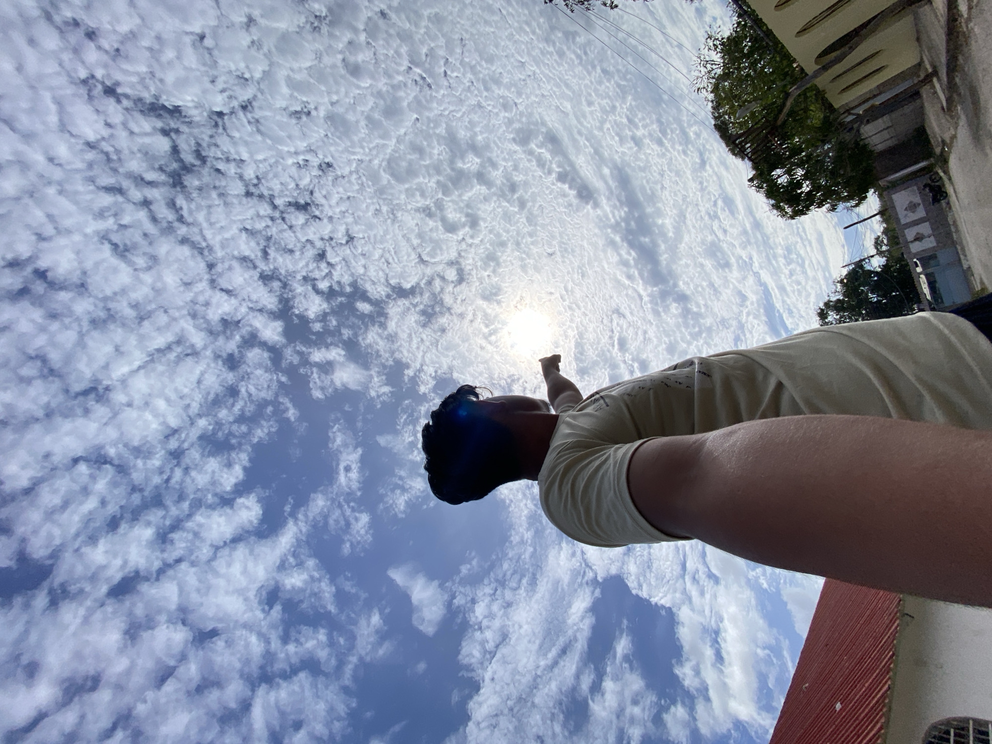 A low-angle shot features a person reaching towards the sun under a sky filled with altocumulus clouds. The sun is positioned near the fingertip of the person's extended hand. The person has dark hair and is wearing a light-colored t-shirt. In the background, buildings with red corrugated roofs and trees are partially visible. The location is Juan Griego, Venezuela.