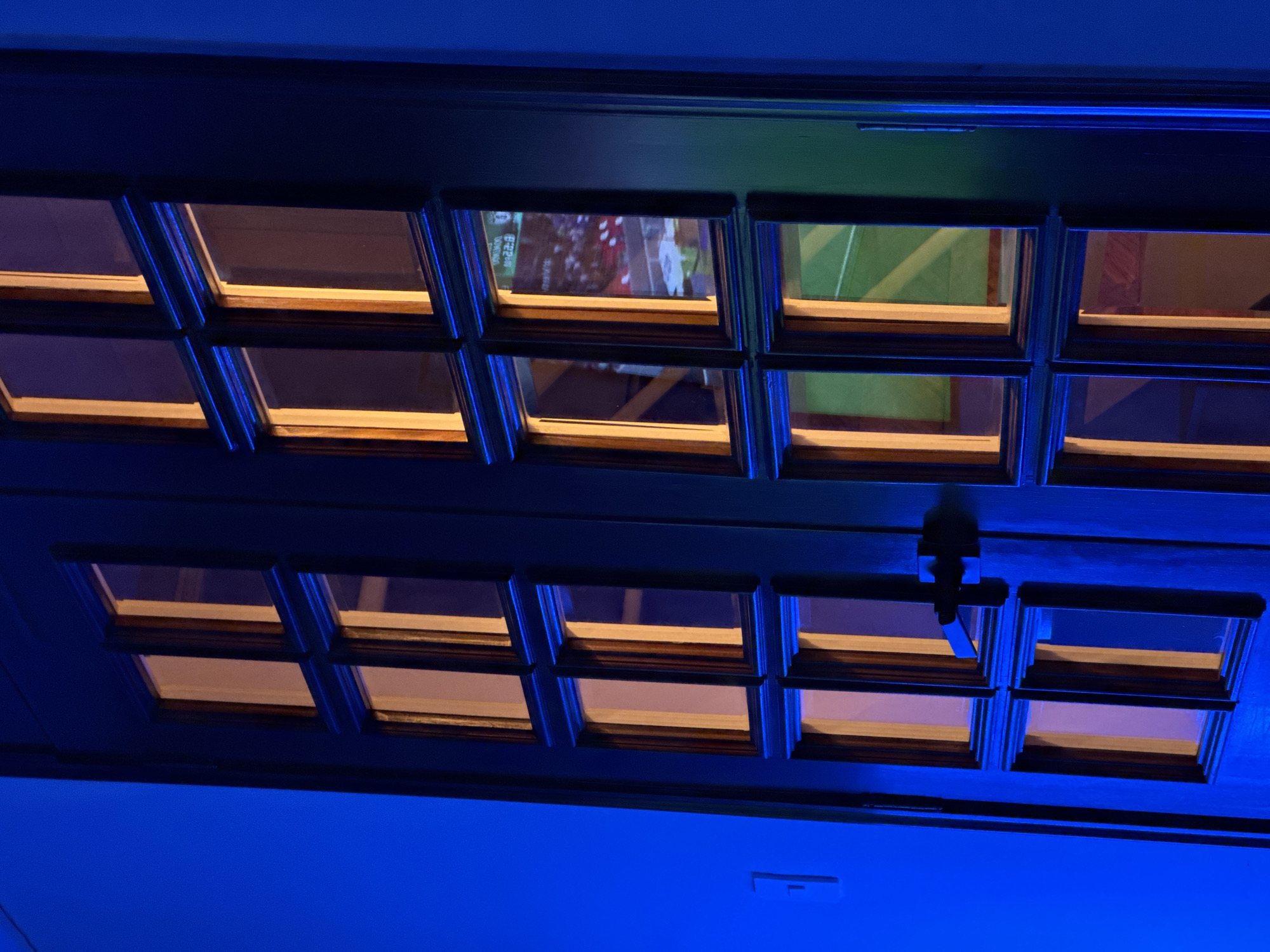 A low-angle shot reveals a dark, likely wooden, door or panel fitted with multiple small, rectangular glass windows. The windows are arranged in a grid-like pattern, and some of them reflect light, while one appears to reflect an image of a sports game. A handle is centrally positioned on the door. The door is surrounded by a bright blue ambient light. The location is Mexico City, Mexico.