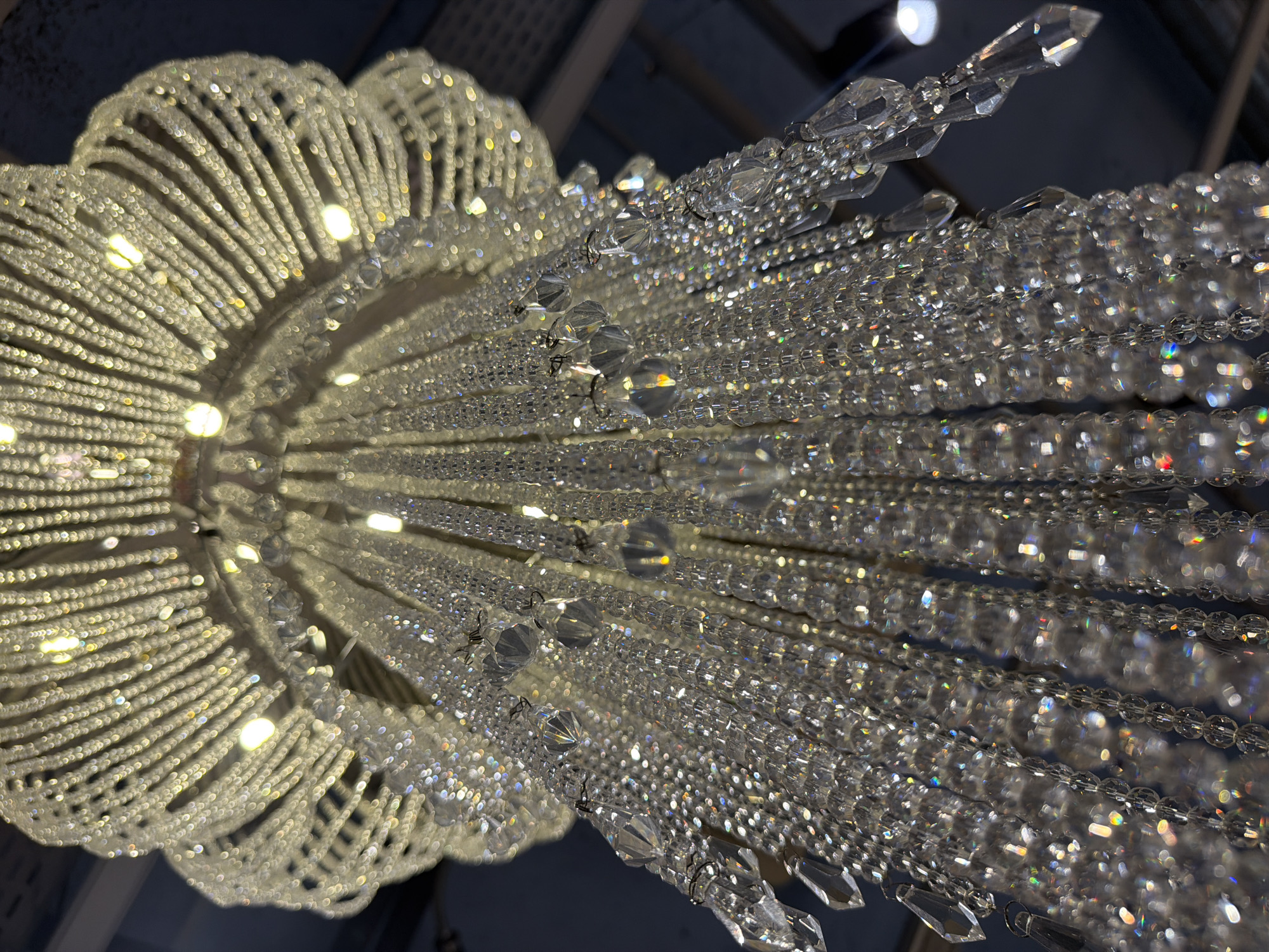 The image depicts a crystal chandelier viewed from below. The chandelier features a circular, flower-like arrangement of lights and crystal strands. Crystal strands cascade downwards, sparkling and reflecting light. The background is dark, suggesting the chandelier is in a dimly lit room. The location is unspecified.