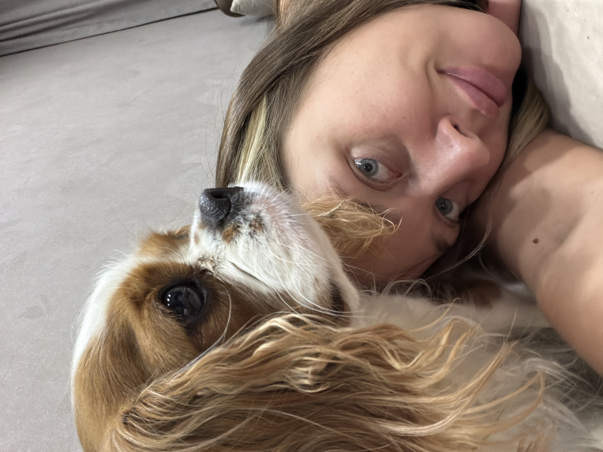 The image displays a close-up, horizontal shot of a female-presenting individual and a dog in an indoor setting. The individual, positioned in the lower right, has light brown hair with blonde streaks, light blue eyes, and light skin. She is lying on a light-colored surface, resting her head, and looking directly towards the camera with a subtle smile. Her left shoulder and upper arm, bearing a small mole, are visible. Adjacent to her head and occupying the left side of the frame is a Cavalier King Charles Spaniel, distinguished by its Blenheim (chestnut and white) coloring, long wavy ears, and a black nose. The dog's head is resting near the person's, and it is looking slightly to the right, away from the camera. The background is predominantly a plain, light gray wall, with a dark gray curtain or fabric partially visible on the far right edge of the frame. The lighting is soft and ambient. The image depicts a calm and close interaction between the person and the dog. The image provides no discernible information regarding its location in the city of Chaikovsky, Russia, or any specific geographical coordinates.