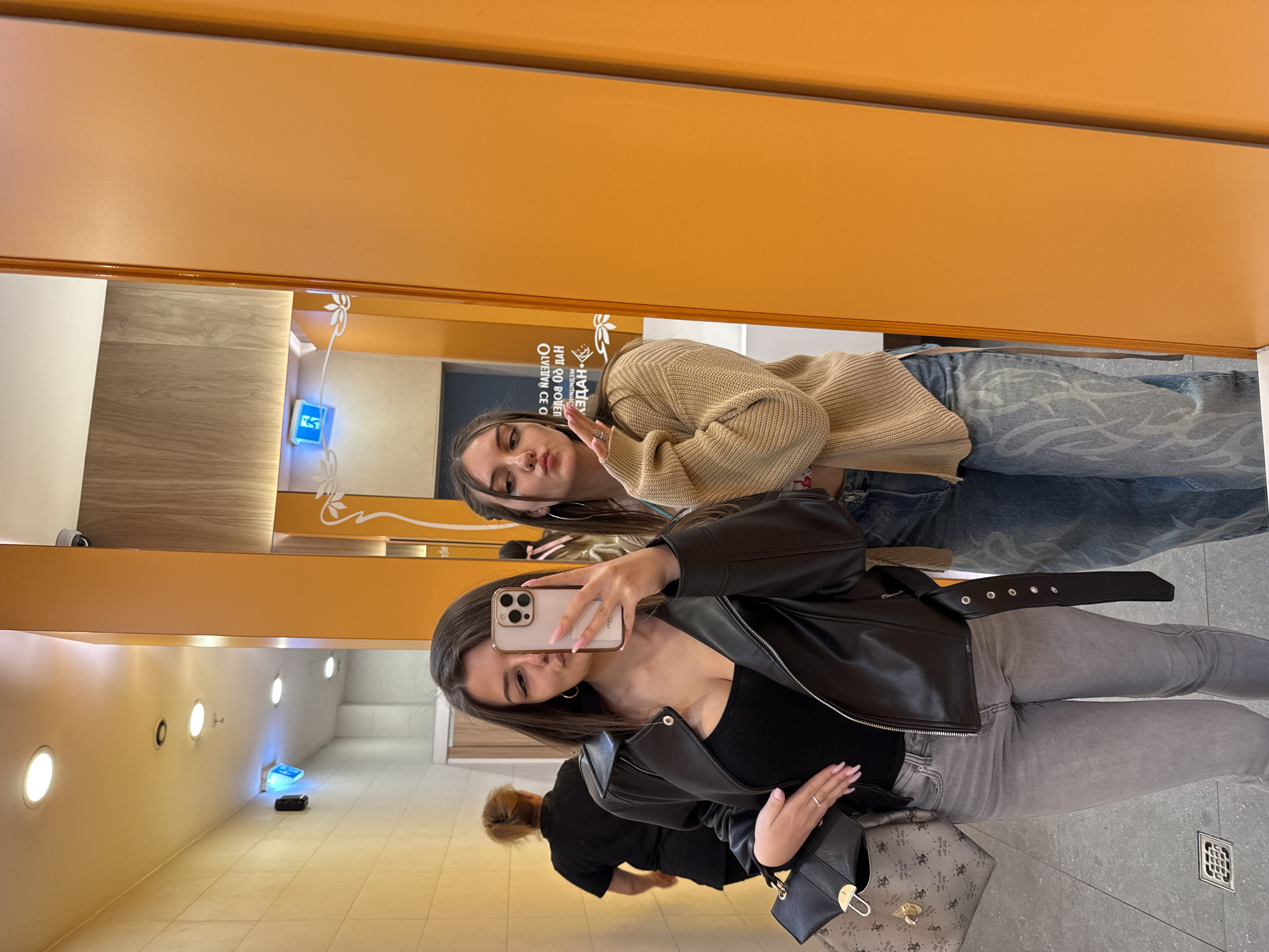 Two young women are taking a selfie in a mirror. One woman wears a black leather jacket over a black top and gray jeans, holding a cell phone. The second woman wears a beige sweater and blue jeans. They appear to be in a public restroom with light yellow walls and tile flooring. The reflection shows other people in the background. A sign written in Bulgarian is visible on the wall. The location is likely Sofia, Bulgaria, based on the language of the sign.