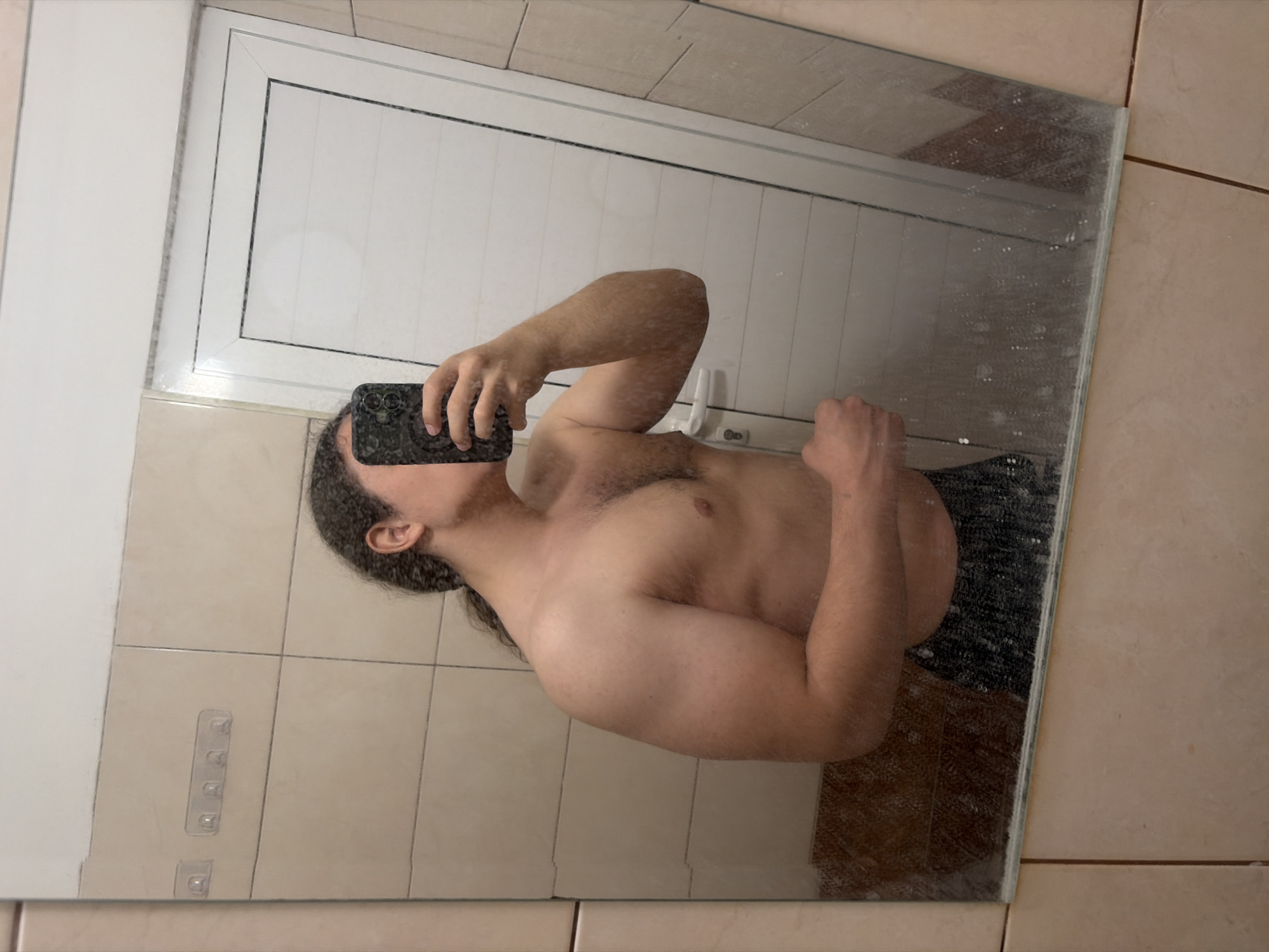 The image, rotated 90 degrees counter-clockwise, displays a mirror selfie of a shirtless person. The individual, seen from the chest up, has long dark hair and some facial hair, holding a dark-colored smartphone to capture their reflection. Their right arm is flexed, showcasing developed bicep muscles. Visible chest hair is present. The mirror itself appears somewhat streaky or dusty.
Reflected in the mirror, the background reveals light-colored square wall tiles, characteristic of an indoor setting, possibly a bathroom or utility room. In the upper part of the reflection, a white framed structure, resembling a door or window frame, is visible. Towards the lower-left of the reflection, a clear plastic strip with multiple hooks is mounted on the tiled wall. Dark clothing or a towel is faintly reflected around the person's lower torso. No visible text is present in the image. The time of day is not discernible.