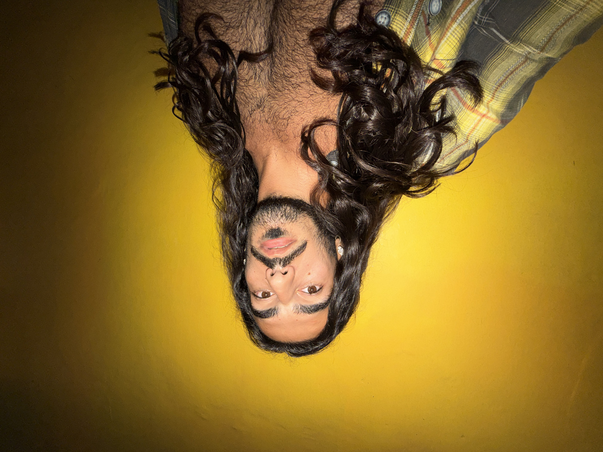 In a selfie taken against a bright yellow wall, a person with long, dark, curly hair and a dark mustache and beard poses shirtless. They are wearing a plaid shirt partially unbuttoned, revealing a chest covered in dark hair. The background is a gradient of yellow, darkening to brown on the right side. No specific location in North Kuta, Indonesia, can be determined from the image.