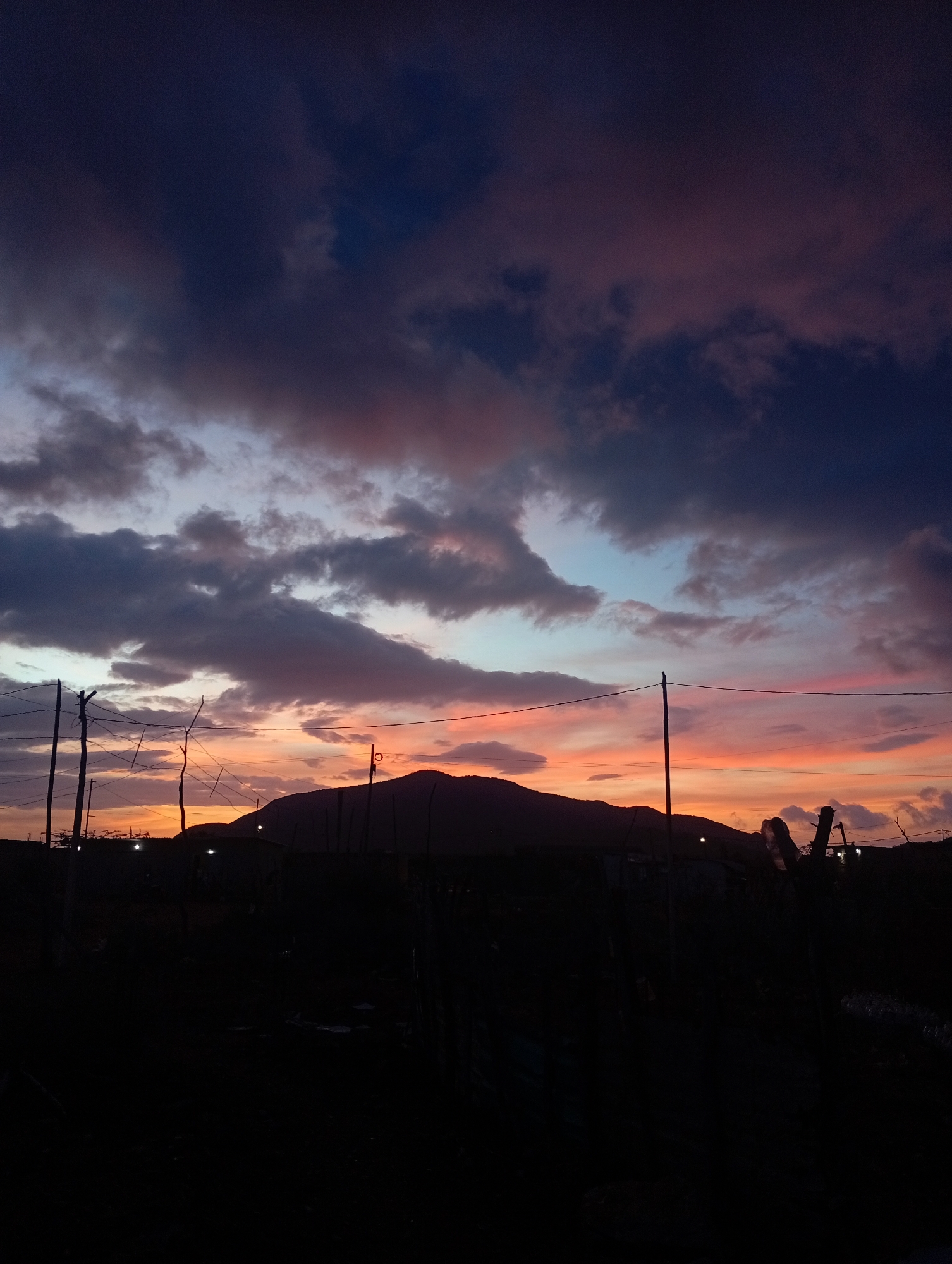 The image shows a silhouetted landscape at dusk, likely in Altagracia, Venezuela. The sky is filled with dramatic clouds painted in hues of deep blue, purple, pink, and orange. A mountain range is visible in the distance, also silhouetted against the vibrant sky. In the foreground, there are dark silhouettes of utility poles, wires, and some vegetation. The lower part of the image is very dark, obscuring details of the immediate ground and any structures or people present. The scene evokes a sense of twilight in a semi-rural or undeveloped area.