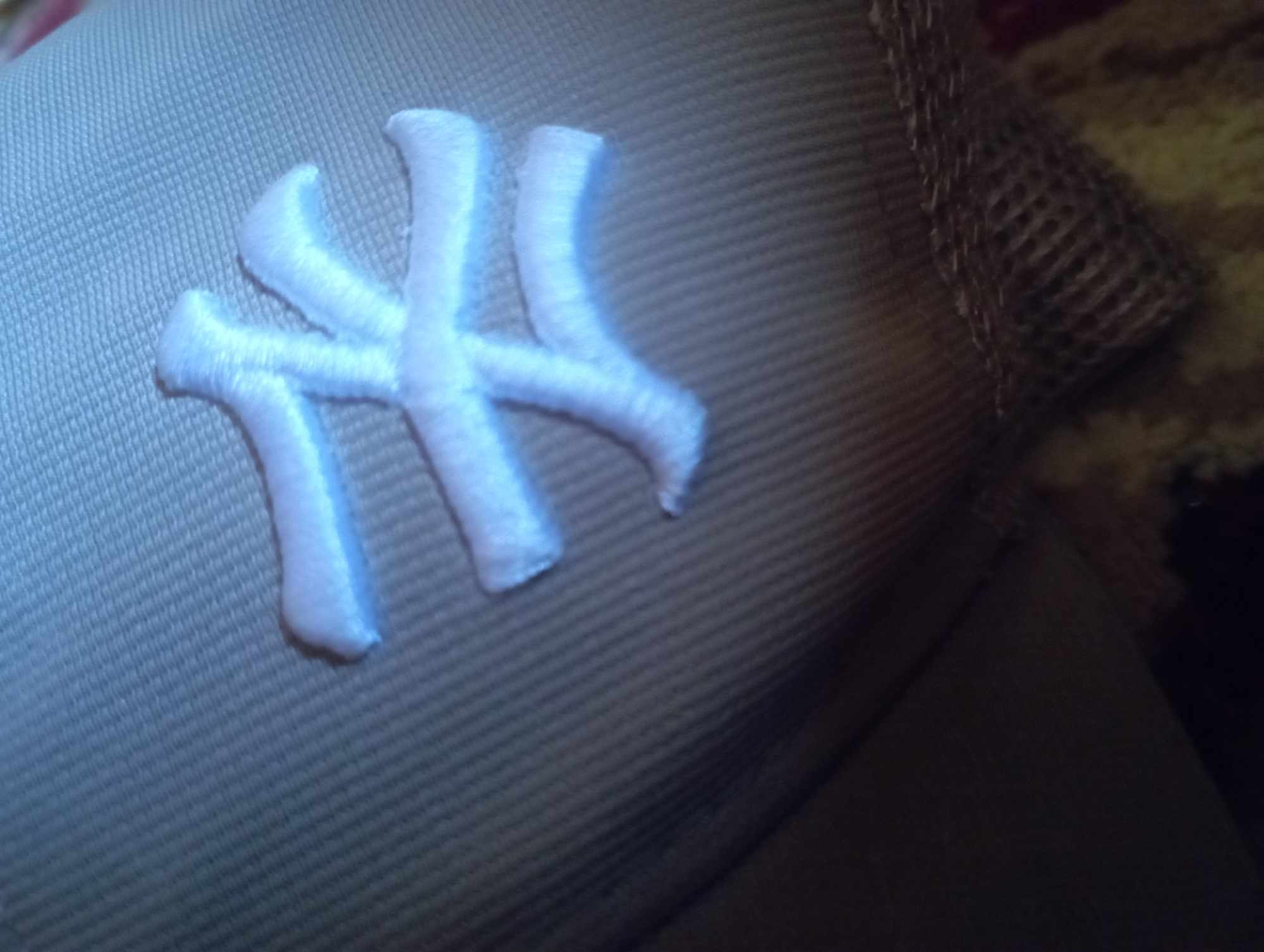 The image shows a close-up view of the embroidered logo on a baseball cap. The logo is white and appears to be the iconic "NY" logo of the New York Yankees. The cap is a light tan color and has a mesh panel on the side, typical of many baseball caps. The lighting is somewhat dim, and the background is out of focus, appearing to be a textured surface, possibly fabric. There is no discernible information about the location in Ezbet Michel Hamawi, Egypt, as the image solely focuses on the cap.