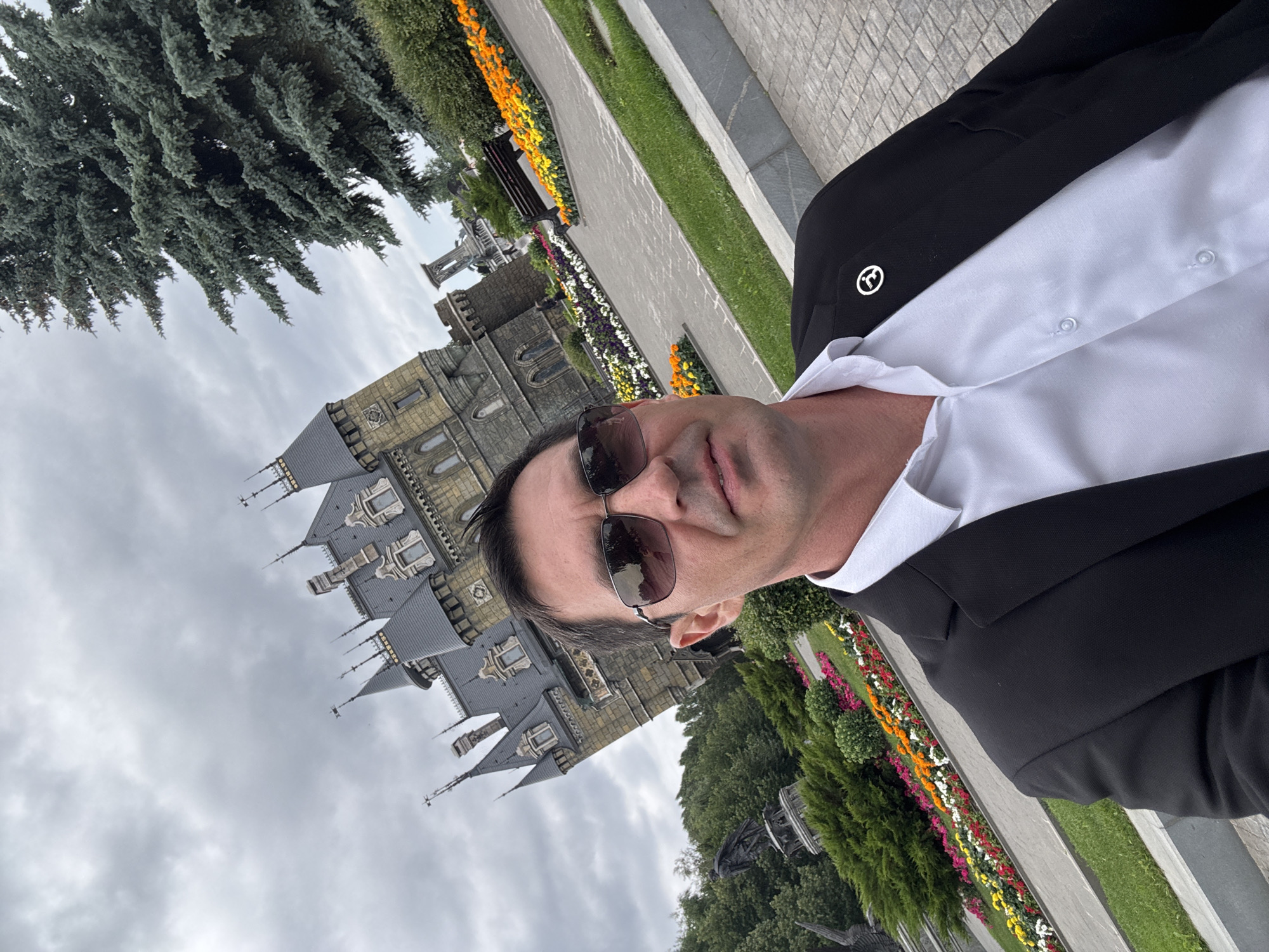 A man in a black suit and white shirt takes a selfie in front of the Garibaldi Castle in Khryashchevka, Russia. The castle, with its distinctive Gothic architecture, is partially visible in the background, along with a large evergreen tree. The scene is set on a cloudy day. The man is wearing sunglasses, and a small badge is pinned to his jacket. In front of the castle, there are flowerbeds with colorful flowers and a paved walkway. A park bench is visible to the right of the castle.