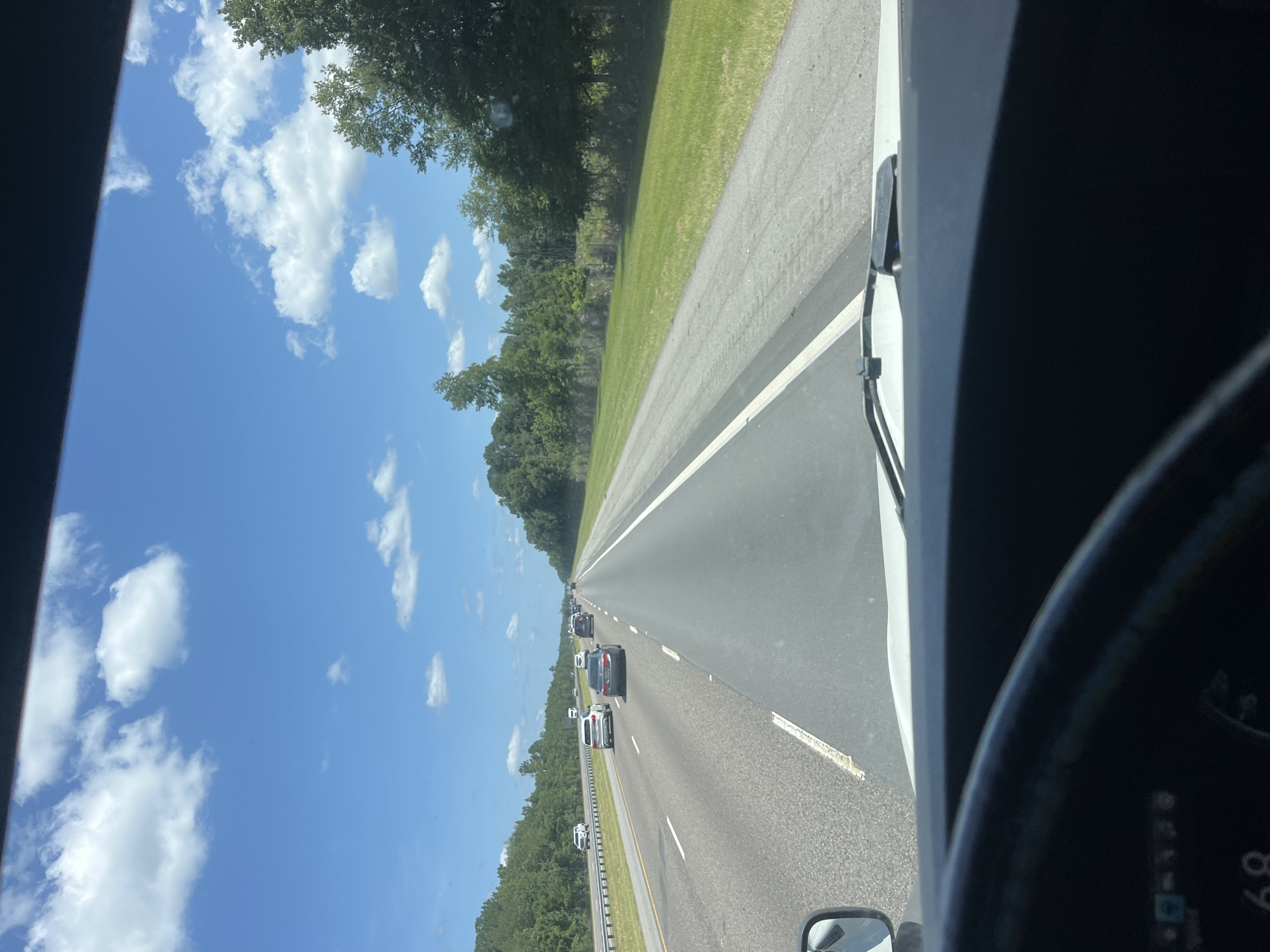 The image is taken from the interior of a vehicle, likely a truck, as evidenced by the steering wheel and dashboard in the foreground and a side mirror on the left. The vehicle is on a highway, with multiple lanes of traffic visible. Other vehicles, including cars and SUVs, are driving on the highway. The environment is a sunny day with a clear blue sky and scattered white clouds. Lush green trees and grass line the sides of the highway, suggesting a rural or suburban setting. The road appears to be in good condition, with clear lane markings. The time of day appears to be daytime, based on the bright sunlight and shadows. There is no visible indication of the specific location in the city of Riceboro, United States, in the image itself.