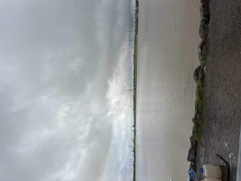The scene, located in Oumazza, Morocco, depicts an expansive body of murky, brownish-gray water under a uniformly overcast sky. The sky is filled with dense, light gray clouds, showing no visible sunlight. The water surface exhibits subtle ripples, extending from the immediate foreground to the distant horizon. Along the far shoreline, a low, flat landmass is visible, punctuated by numerous tall, metal lattice-work electricity transmission towers, some partially obscured by a hazy atmosphere. Scattered green vegetation or trees are present along this distant bank. In the immediate foreground, a dark, granular ground surface, likely gravel or dirt, runs horizontally across the bottom of the frame. This ground abuts a low embankment constructed of dark, irregular rocks and interspersed with small patches of green grass and weeds. On the far left of the foreground, two light-colored, possibly plastic, jugs with blue caps are visible, resting on a dark, flat surface, alongside a crumpled blue plastic bag. A white, flat object, potentially a table edge, is partially visible at the extreme bottom left corner. No individuals or active interactions are observed in the image.