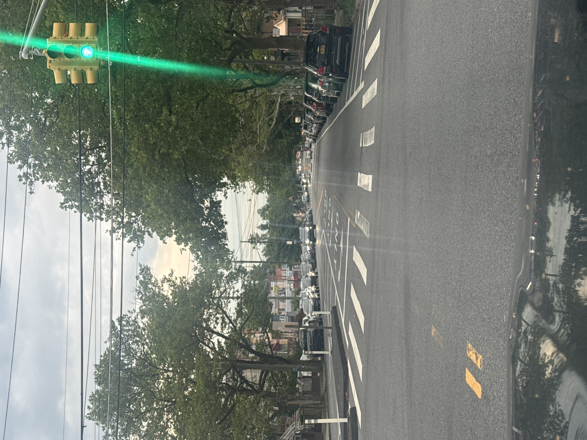 The image captures a street view in New York City, USA, characterized by a cloudy sky overhead. The scene is dominated by a paved road with clearly marked crosswalks and lane dividers. Several cars are parked along the right side of the road, with more vehicles visible further down the street. Trees line both sides of the road, their green foliage creating a canopy effect in some areas. A traffic light is prominently displayed on the right, showing a green signal. The overall impression is of a typical urban residential street.