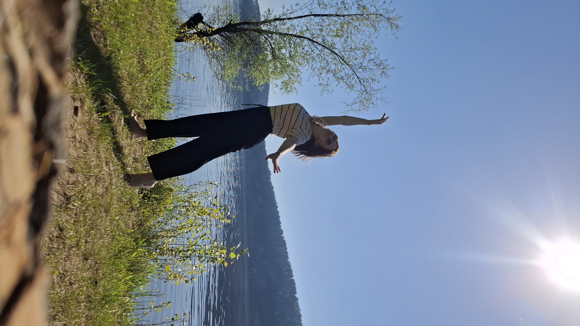 A young woman stands barefoot on a grassy bank by a body of water, possibly in the city of Noviy, Russia. She wears dark pants and a striped top, with her arms outstretched. A tree with light green leaves is behind her. In the background, a forested hill is visible across the water. The sky is clear blue with the sun shining brightly, creating a lens flare. The perspective of the image is rotated, giving the impression that the scene is tilted.