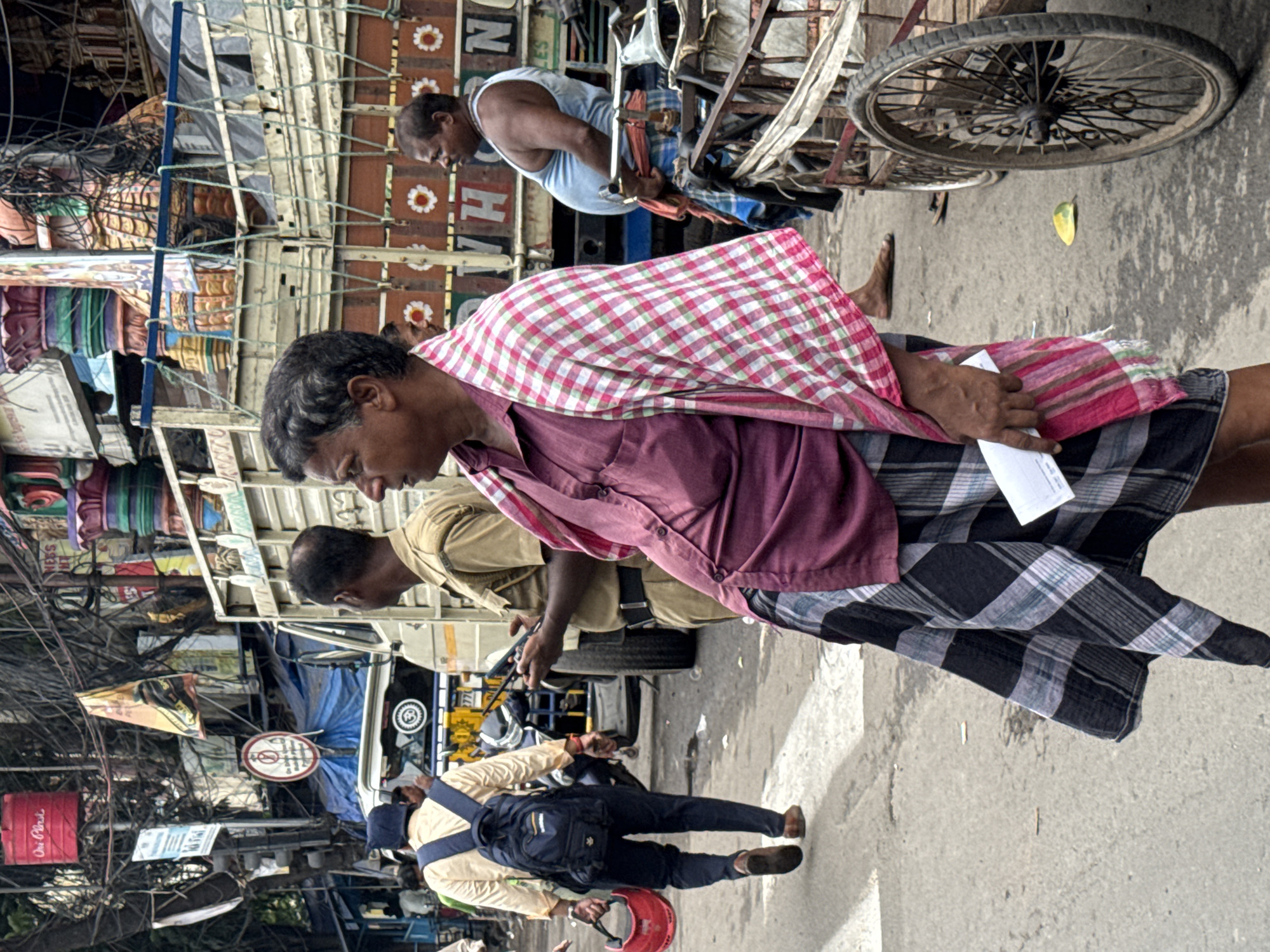 In Kolkata, India, a busy street scene unfolds with several men and a truck. A man in the foreground, wearing a maroon shirt, a checkered lungi, and a pink and white checkered cloth draped over his shoulder, holds a piece of paper. To his left, another man in a tan uniform and a backpack walks away, carrying a red helmet. Behind them, a large truck is parked, adorned with colorful decorations. Another man in a light blue tank top stands near the truck, and a third man in a tan uniform is visible. A pushcart with large wheels is positioned on the right side of the frame. The street appears to be a market area, with overhead wires and street signs adding to the urban atmosphere.