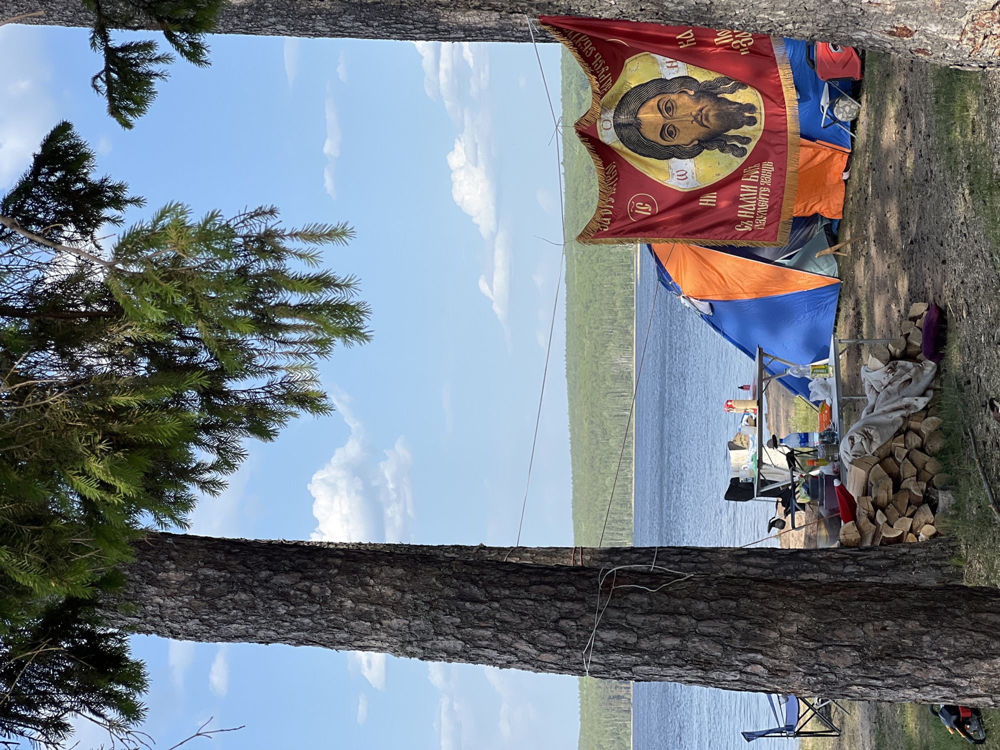 On the shores of a body of water, likely the Kama Reservoir, near Chaikovsky, Russia, a campsite is established. Visible between two tree trunks, a red flag bearing an Orthodox Christian icon hangs above tents of various colors, including blue and orange. A dock extends into the water. A pile of firewood rests near a table laden with supplies. A blue camping chair sits on the grass. The sky is blue with scattered clouds.