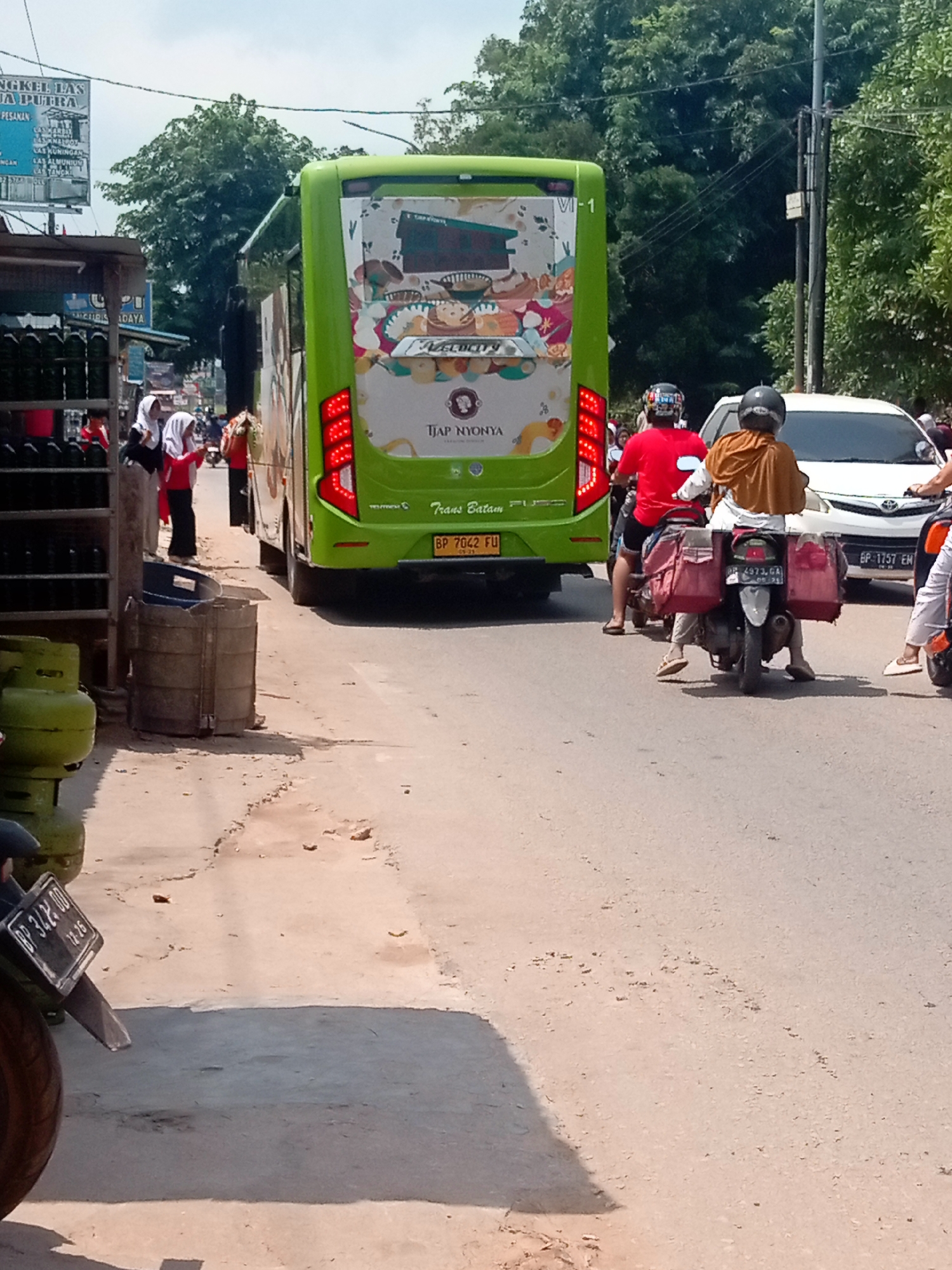 This is a street-level, full shot image of a busy street in Batam, Indonesia, on a sunny day. In the foreground, a motorcycle is parked on the side of the road, with its rear tire and license plate visible. The license plate reads "BP 3424 DD". Further down the road, a green bus with "VI-1" and "TJAP NYONYA" written on its back is prominent. The bus has decorative graphics on its rear. To the left of the bus, a white car is also visible. Several motorcycles with riders are also present on the road, with one rider in a red shirt and another in an orange hijab. People in religious attire, including two women in hijabs, are seen standing by a stall on the left side of the image. The environment includes trees, power lines, and some market stalls. The road surface is asphalt with some dirt and debris.