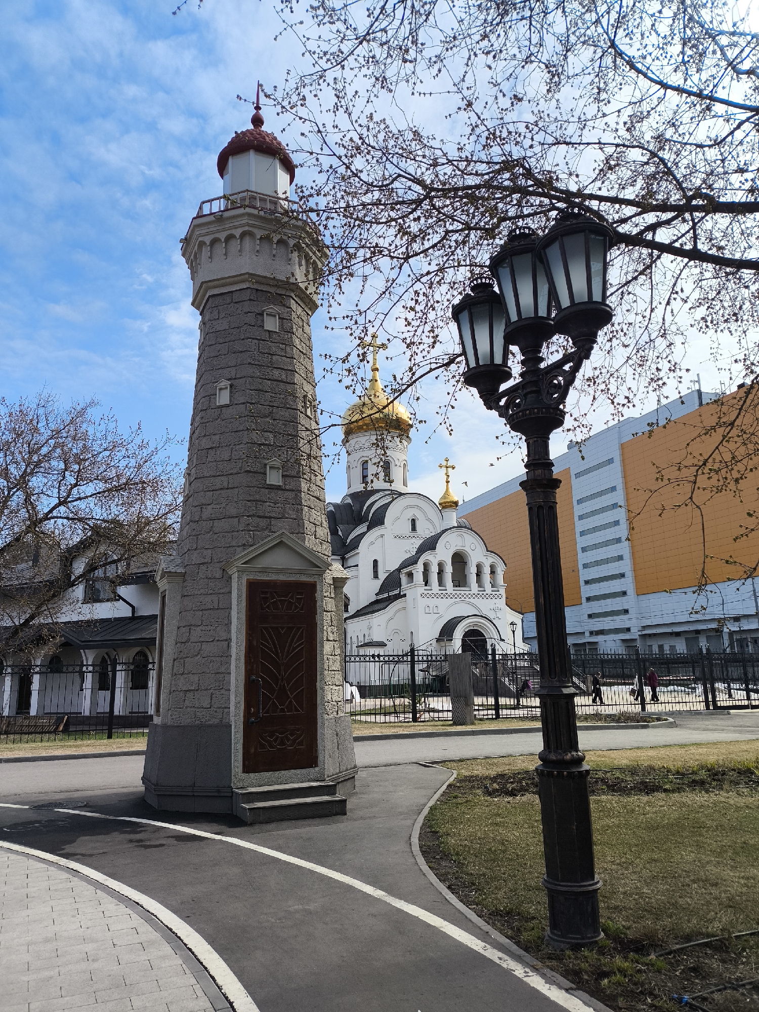 The image depicts a cityscape in Moscow, Russia, featuring a lighthouse-like structure with a red roof, a Russian Orthodox church with golden domes, and a lamppost. The lighthouse stands on a paved area with a dark door. The church is white with arched windows and a golden dome topped with a cross, situated behind a black metal fence. To the right of the church, a large modern building with an orange facade is visible. A black lamppost with three lanterns stands to the right of the lighthouse. Bare tree branches frame the scene. Several people are walking in the background near the fence. The sky is light blue with clouds.