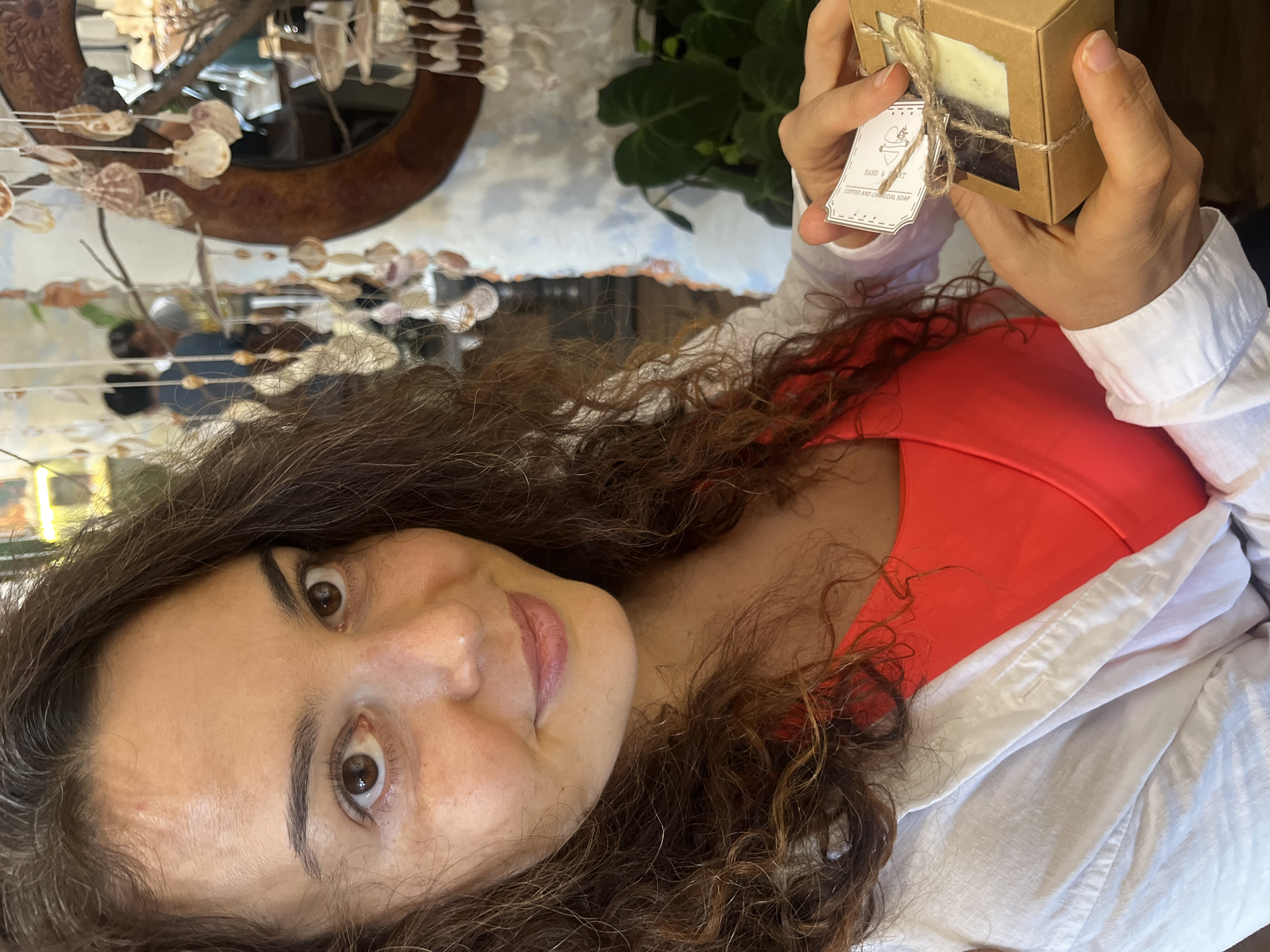 A woman with curly brown hair and brown eyes is holding a small, brown cardboard box containing two bars of soap. The box is tied with twine, and a tag with text and an illustration is attached. She is wearing a bright orange tank top and a white open button-down shirt. The background appears to be a shop or market, with shelves, plants, and decorative seashell hangings visible. The lighting suggests it is daytime, possibly indoors.
