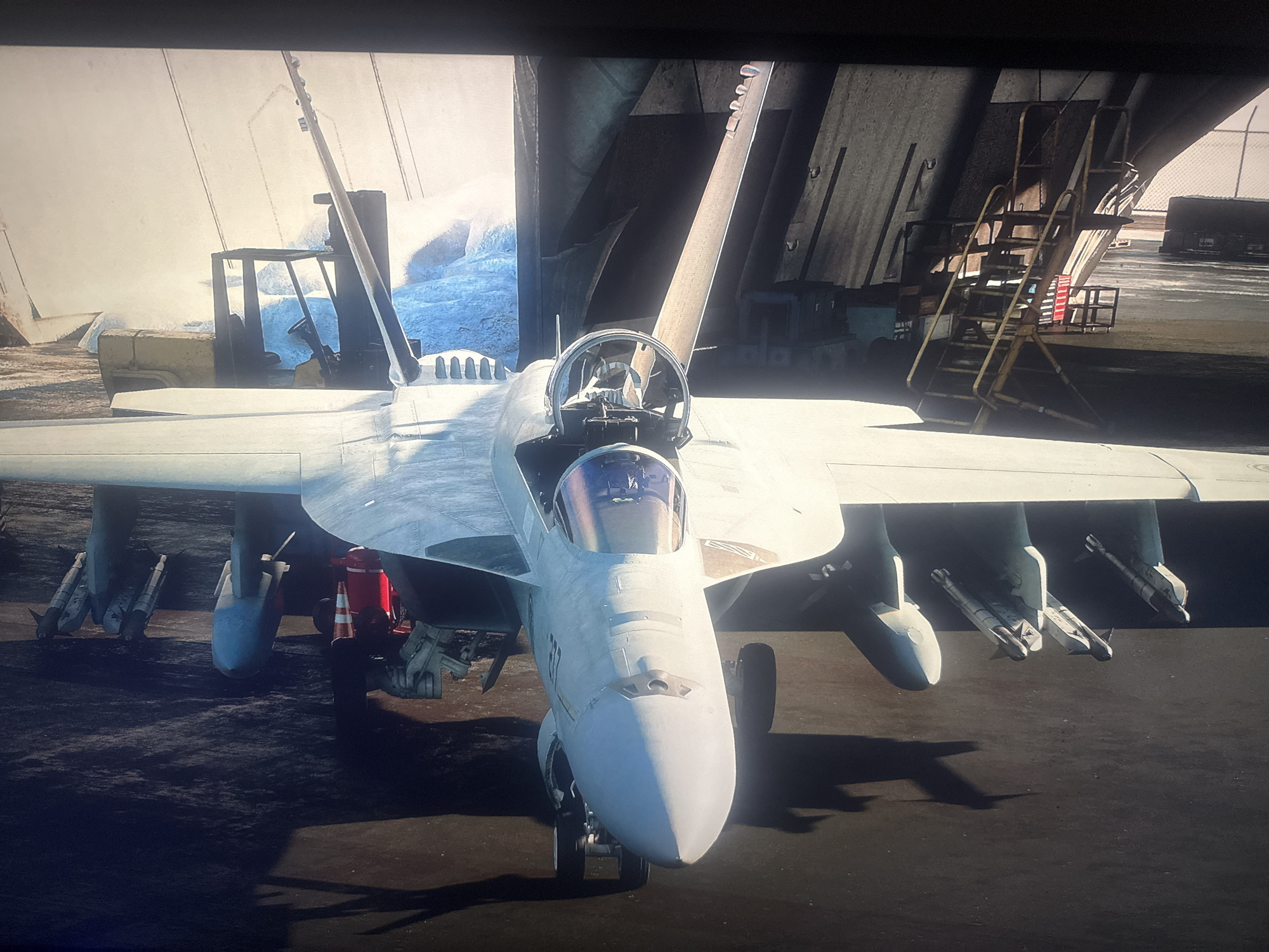 An overhead, front-facing view captures a fighter jet parked on an aircraft carrier. The jet, painted in a light gray, is equipped with missiles on its wings. Behind the jet, the carrier's structure and a yellow ladder are visible. The scene appears to be set within a hangar, with hints of outdoor light in the background. This image does not depict the city of Herzliya, Israel.
