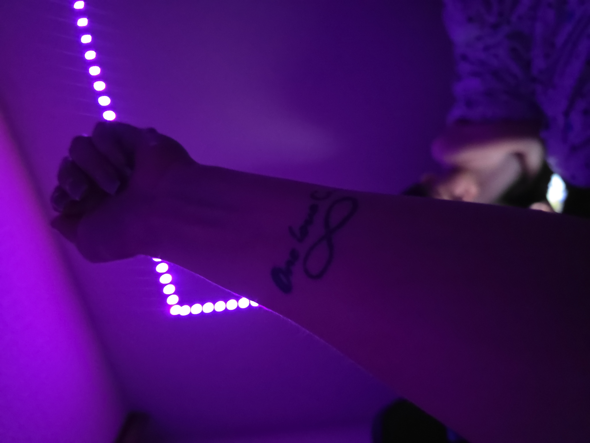 The image depicts an indoor scene bathed entirely in deep violet and purple artificial light, likely originating from LED strip lights, some of which are visible as a string of bright, individual circular emitters along the upper and right edges of the frame. In the foreground, a fair-skinned human forearm and a clenched fist are raised, extending from the lower-left towards the upper-right. The inner forearm displays a black ink tattoo featuring the cursive text "One love" positioned above a black ink infinity symbol. In the blurred background, towards the lower-right, a second individual is partially visible, reclining under a textured, purple-hued blanket or duvet. A small, bright, rectangular light source, consistent with a mobile phone screen, is visible near their hands. The walls appear plain, reflecting the pervasive purple light. A dark, indistinct shape is present in the upper-left corner, possibly a ceiling fixture. The specific elements within the image do not provide direct visual cues to identify the city of Minsk, Belarus; however, the scene is situated within a location in Minsk, Belarus, as per the prompt's context.