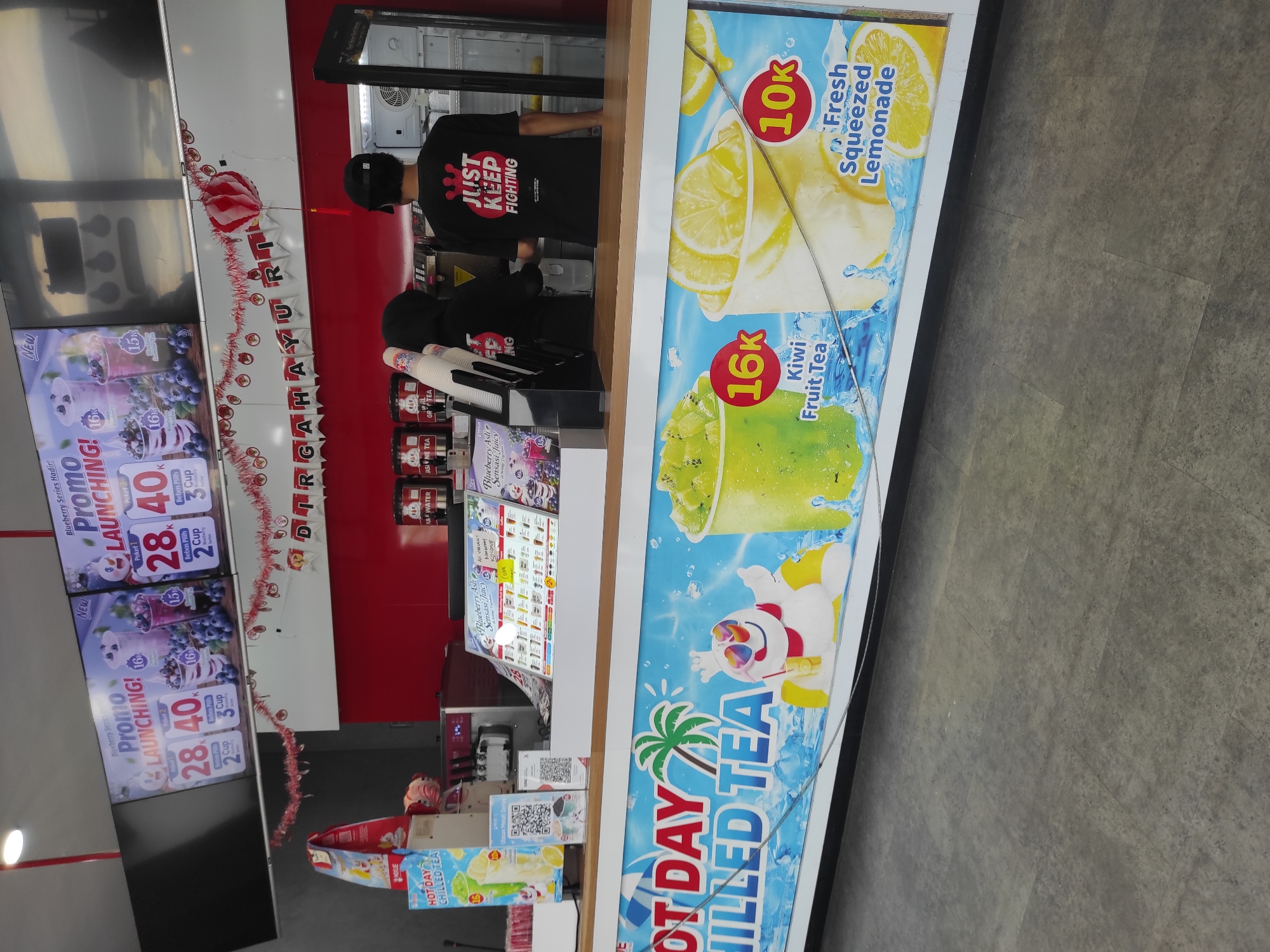 The image shows a storefront for a beverage shop, likely in Pleret, Indonesia, based on contextual clues. A large, colorful banner along the bottom of the counter advertises "HOT DAY CHILLED TEA" with graphics of tropical drinks and a cartoon character. Behind the counter, two employees, one wearing a black t-shirt with the text "JUST KEEP FIGHTING" and the other a black hoodie, are interacting. Above them, promotional posters display beverage specials, including "Blueberry Series" launches with prices in Indonesian Rupiah (e.g., 28,000 and 40,000). A red and white striped decoration is hung across the top of the display area. The environment appears to be an indoor retail space with tiled flooring and a clean, well-lit counter.