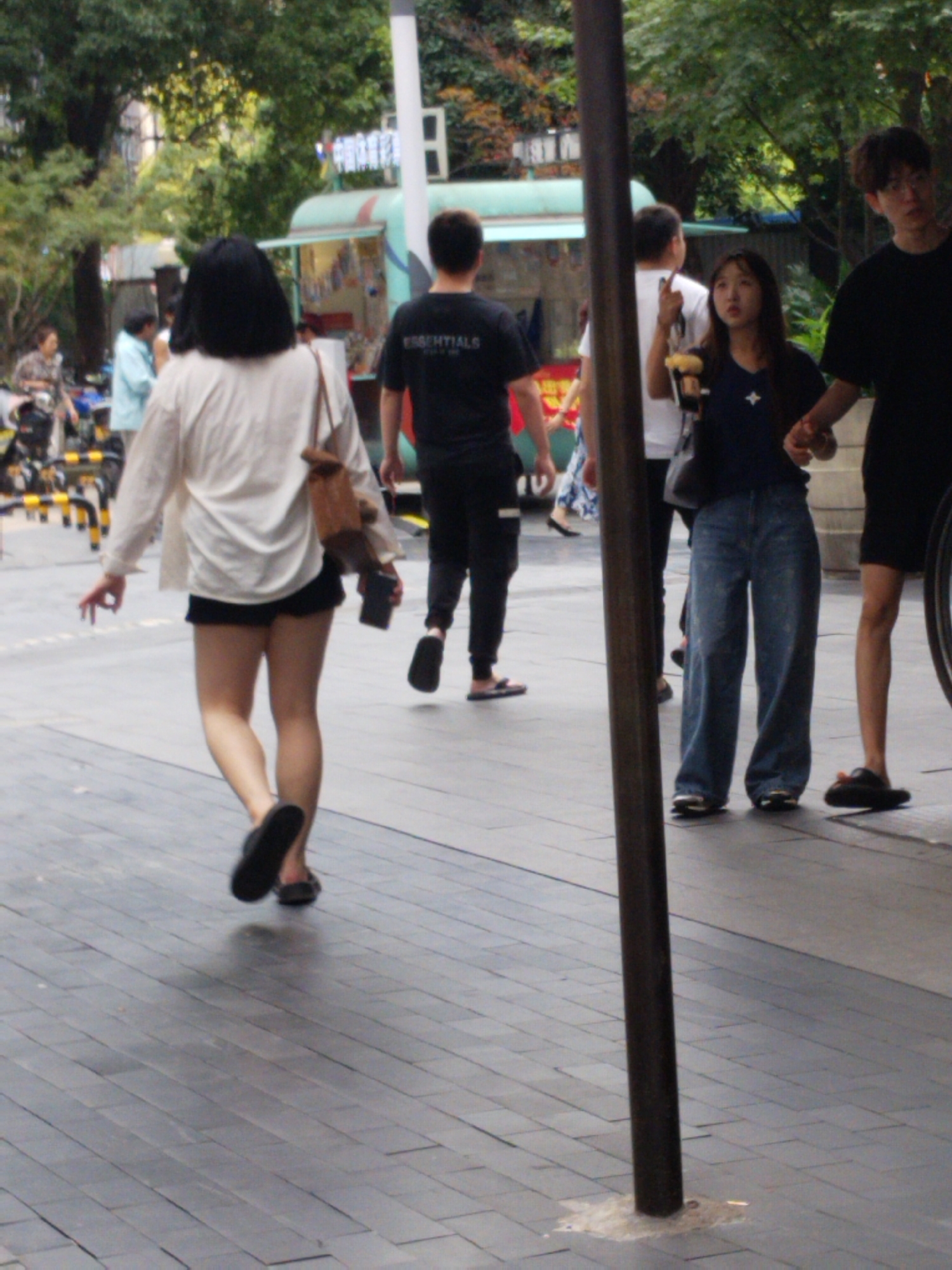 A street scene in Yuzhong District, China shows pedestrians on a paved sidewalk. A woman with dark hair, wearing a white shirt and dark shorts, walks away from the viewer. A man in a black t-shirt with the word "ESSENTIALS" on the back is walking in the same direction as the woman. A young couple stands on the sidewalk, holding hands. The woman is holding a drink. Other pedestrians and a vehicle are visible in the background. A brown pole is visible in the foreground.
