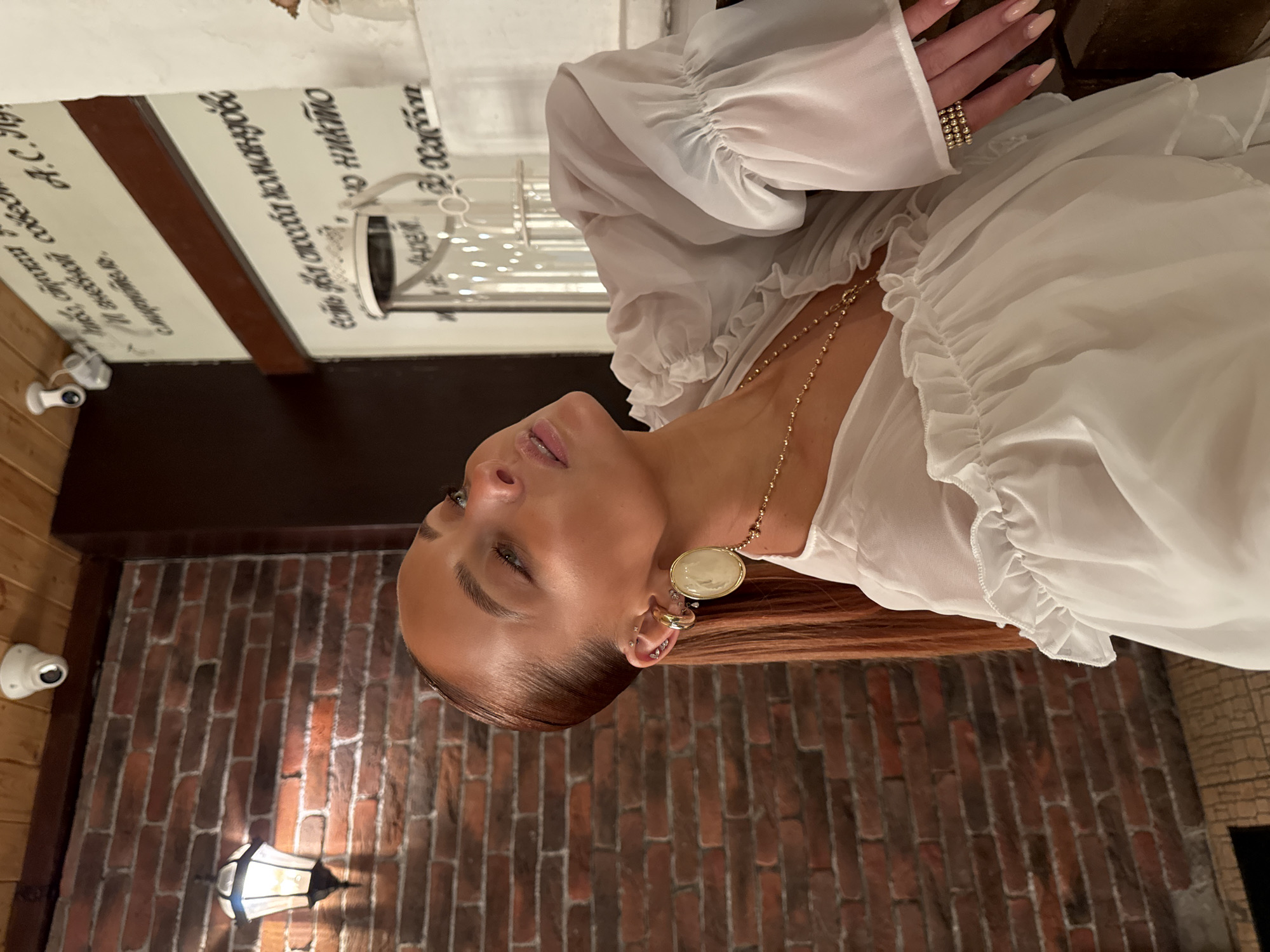 A woman with red hair pulled back in a sleek ponytail is captured in a medium shot, her head tilted back and her gaze directed upwards. She is wearing a sheer white blouse with ruffled sleeves and a ruffled neckline. A gold chain with a circular pendant rests on her chest, and she has multiple rings on her fingers, including a stacked ring on her right hand. She also wears large, oval earrings and multiple ear piercings. The background features a brick wall on the left and a wall with text and a shelf on the right, suggesting an indoor setting. The presence of security cameras on the ceiling indicates this location may be a public or commercial space. The text on the wall appears to be in Cyrillic script, potentially indicating a Russian-speaking environment. While the image offers clues such as Cyrillic script and common architectural elements, it does not contain specific landmarks or details that definitively place the scene in the city of Chaikovsky, Russia.