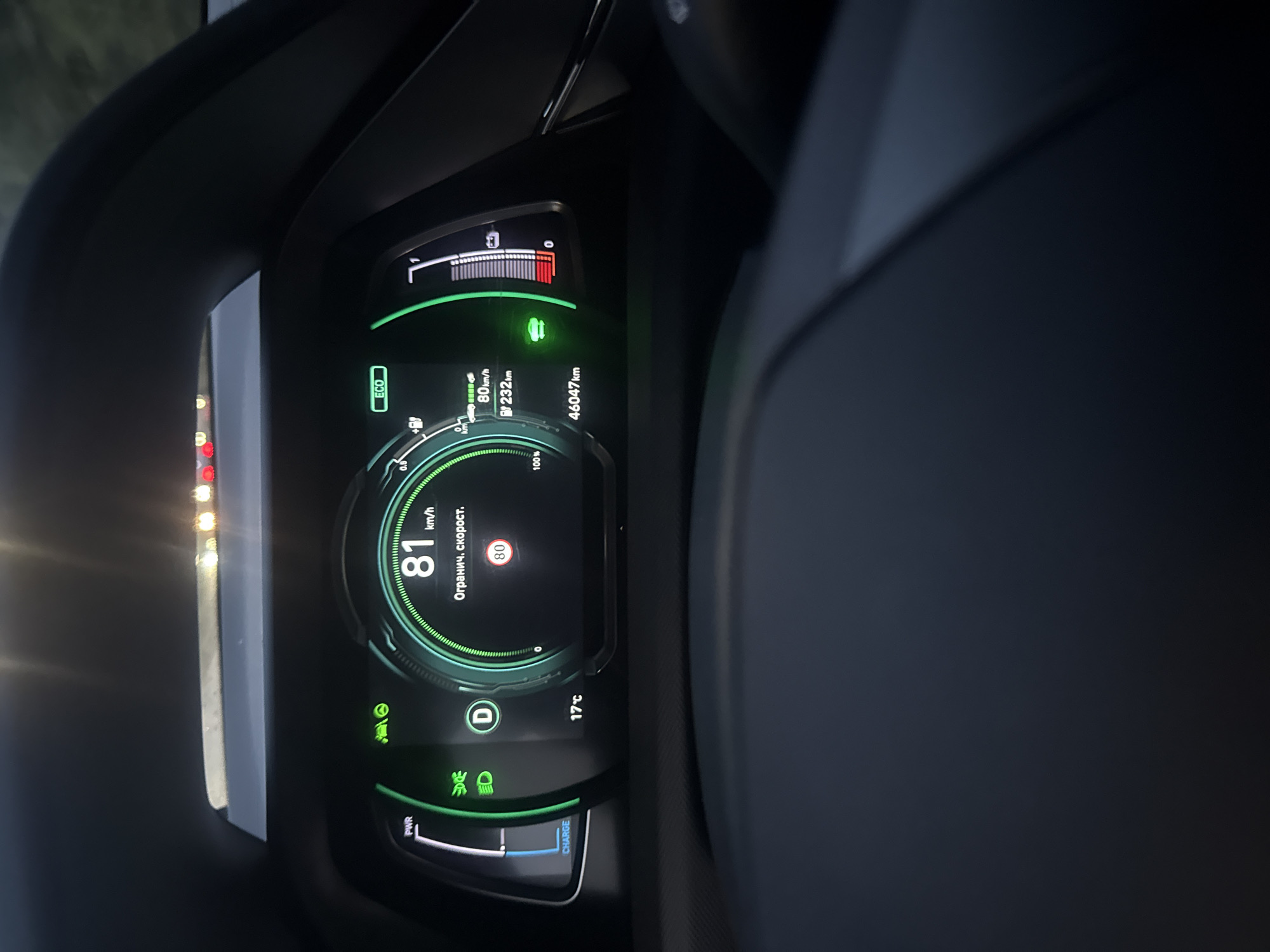 The image shows a close-up view of an electric car's dashboard display at night, likely in Oslo, Norway, given the visible outdoor lighting. The display shows the car's speed at 81 km/h, with a speed limit indicator of 80 km/h, suggesting the car is exceeding the speed limit. The screen also indicates "Огранич. скорост." which translates to "Limited speed." Other information visible includes the ambient temperature of 17°C, the car's odometer reading of 46,047 km, and a battery charge indicator. The "ECO" mode is also activated. The dashboard's interior lighting is illuminated, casting a green glow on the display. The background is dark, indicating it's nighttime, and the car appears to be in motion. The car's steering wheel is partially visible in the foreground.