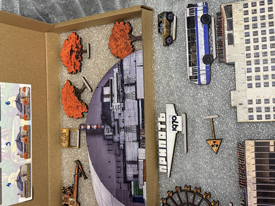 A box contains pieces of a puzzle or model, depicting scenes from Chernobyl or Pripyat. Elements include: the reactor itself, autumnal trees, a rusted car, a blue bus with Cyrillic writing, a Pripyat town sign with "1970," a radiation hazard symbol, a building facade, a Ferris wheel segment, and a crane. A reference card shows numbered scenes for assembly.