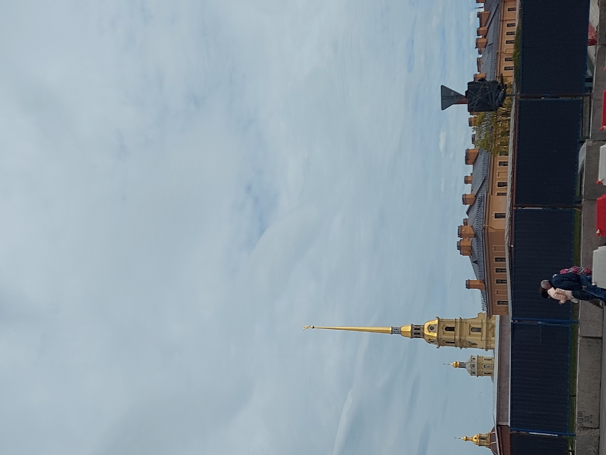 A sideways, eye-level shot shows the Peter and Paul Cathedral tower against a cloudy sky in Saint Petersburg, Russia. The spire of the cathedral is golden and visible near the bottom center of the image. The base of the cathedral, including a section of the wall, is located along the right of the photo. A few people can be seen. A sign of some kind is visible at the top right of the frame.