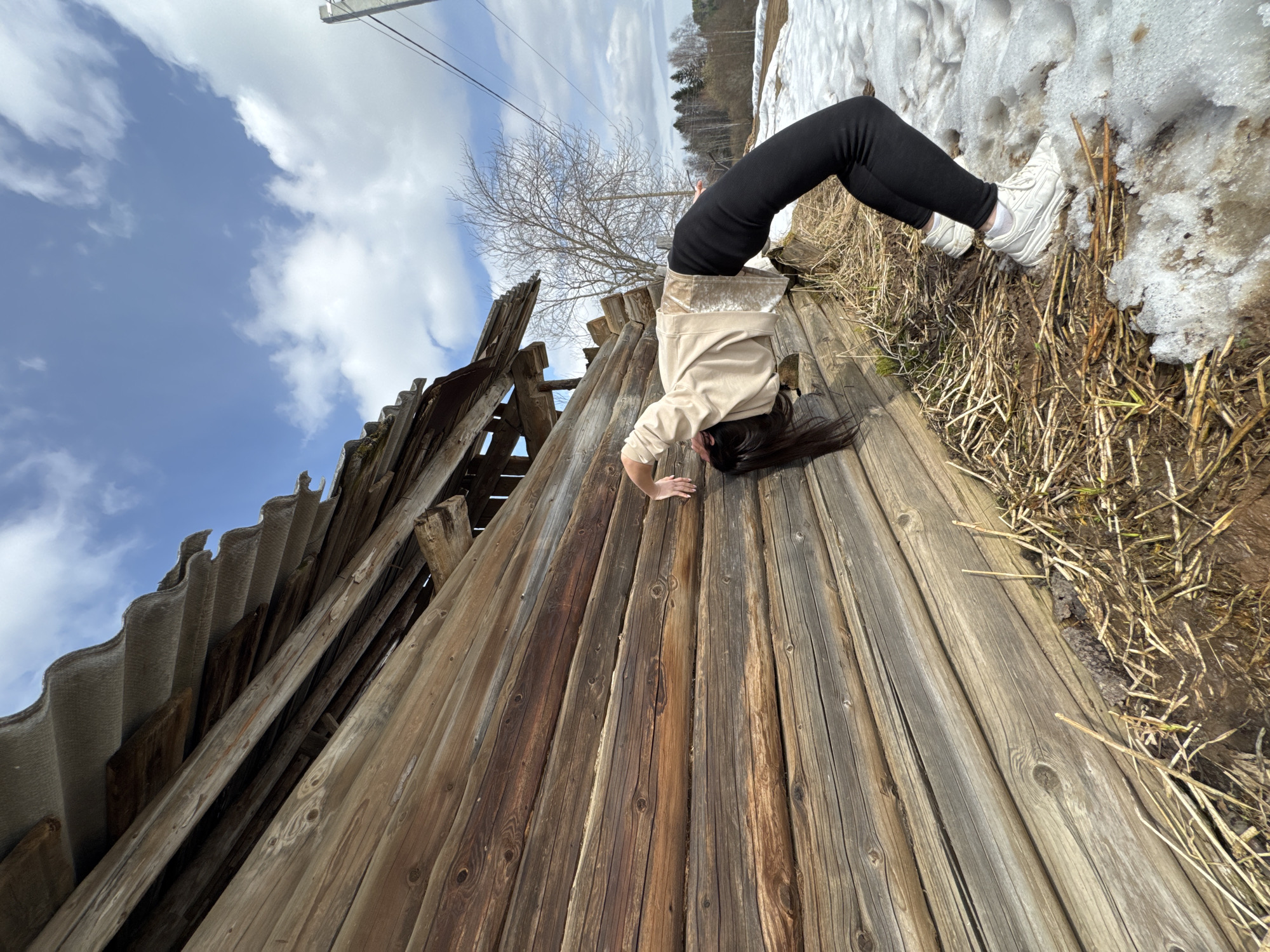 The image shows a woman doing a backbend near a dilapidated wooden structure in Вамсята, Russia. She is wearing black leggings, white sneakers, and a light-colored sweatshirt. Her feet are near a patch of snow and dry vegetation. The structure appears to be an old, weathered building with a corrugated roof and decaying wooden planks. The background shows a partly cloudy sky and distant trees.