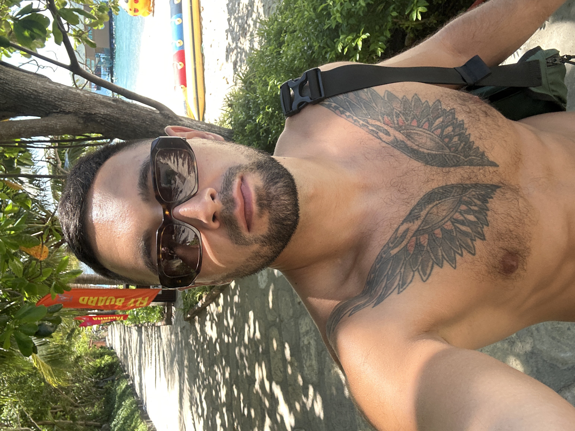 A man with a beard and sunglasses takes a selfie outdoors. He is shirtless, revealing a large tattoo of wings on his chest. He wears sunglasses and a black strap across his shoulder. The background shows a tree-lined path, lush green vegetation, a body of water, and inflatable water toys. A "LIFE GUARD" flag is visible on the left. The location is identified as Bac Nha Trang Ward, Vietnam, with the scene suggesting a beach or resort area. The lighting indicates it is daytime, likely sunny.