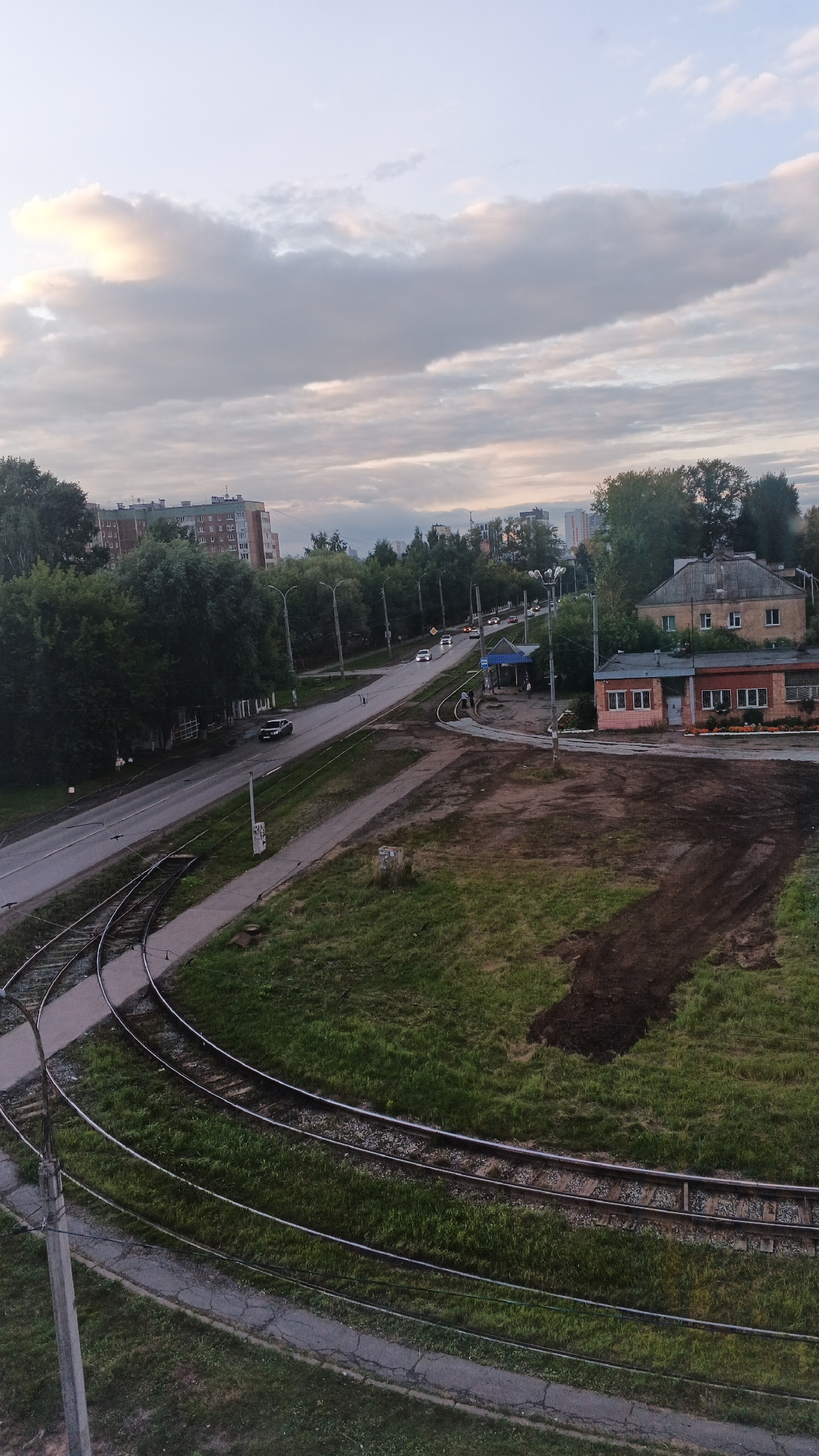 This media file captures an aerial view of a street scene in Izhevsk, Russia, with a focus on urban infrastructure and residential buildings. The scene appears to be set in the late afternoon or early evening, as indicated by the soft, diffused light and the muted colors of the sky, which is filled with scattered clouds.
In the foreground, a complex network of tram tracks curves through a grassy area. Adjacent to the tracks, a paved walkway runs alongside. Further into the frame, a road stretches into the distance, with several cars visible, suggesting moderate traffic. Along the road, streetlights are spaced out, and a bus stop shelter is present.
On the right side of the image, there are two buildings. One is a larger, older-looking building with a pitched roof and multiple windows. Next to it is a smaller, single-story brick building with a light-colored facade and windows. The area between the buildings and the road is a patch of disturbed earth, possibly under construction or recently landscaped.
In the background, a row of apartment buildings and trees form a typical urban landscape. The overall impression is one of a functional, lived-in city environment. There is no visible text in the image. The weather appears to be overcast, contributing to the subdued lighting.
