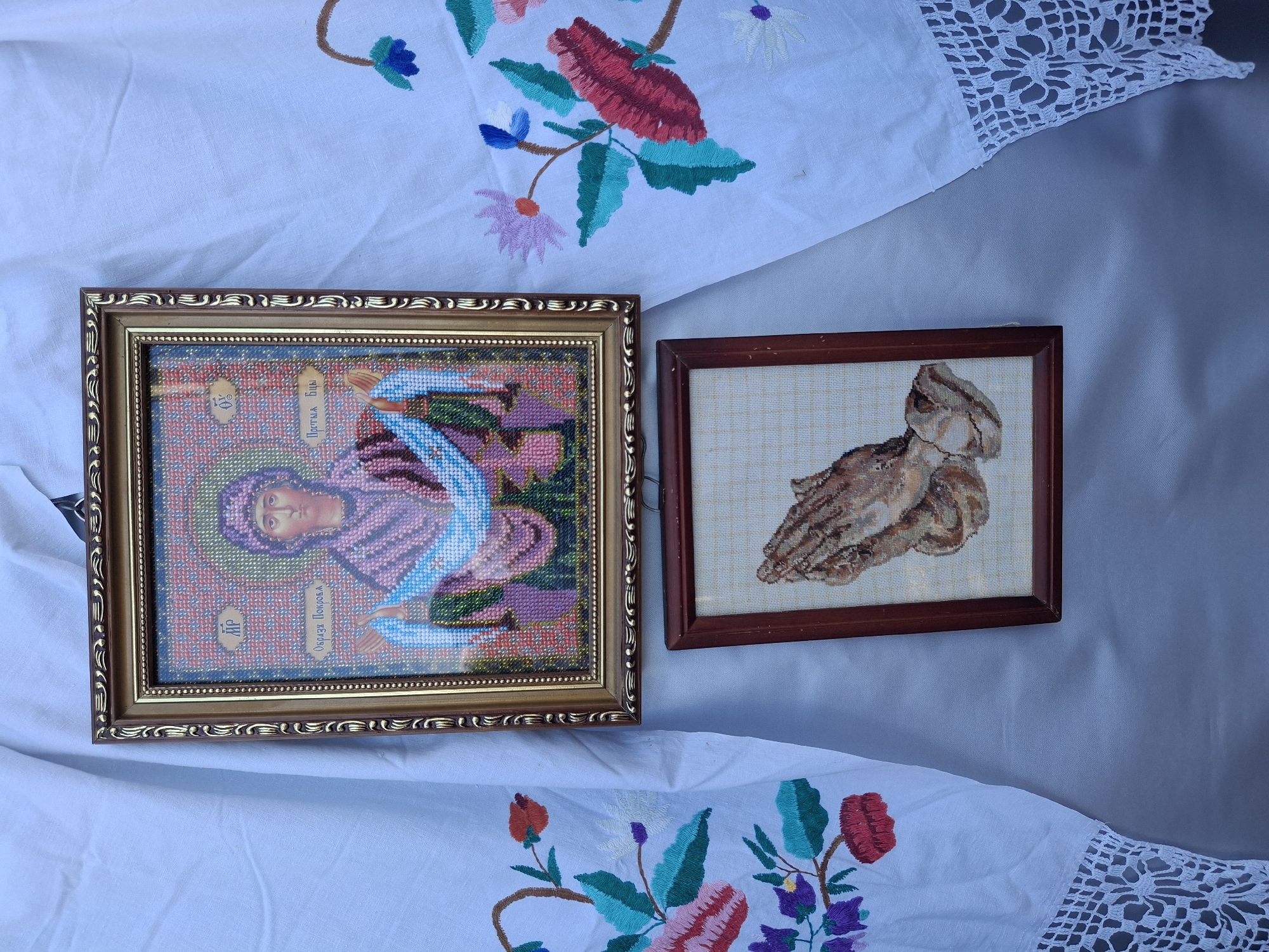 In Roghi, Moldova, two framed embroidered artworks are displayed against a white fabric background. The top artwork, in a gold frame, depicts an icon of the Virgin Mary, rendered in beads. The bottom artwork, in a dark wood frame, shows a close-up of praying hands, also embroidered. The white fabric behind the artworks is decorated with colorful floral embroidery and lace trim along the bottom edge. The scene suggests a cultural or religious display.