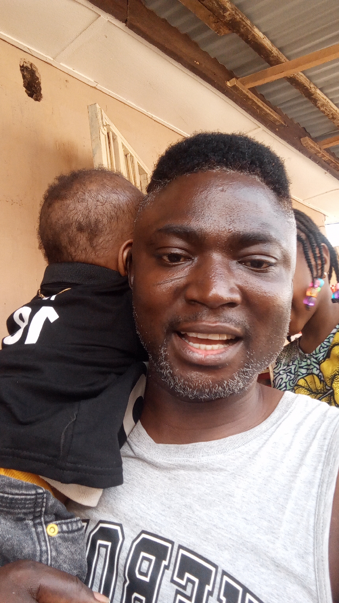 A man is taking a selfie while holding a baby on his shoulder, outside in what appears to be Sabon Tasha, Nigeria. The man is dark-skinned with short, dark hair and a salt-and-pepper beard. He is wearing a grey tank top with a graphic design. The baby is wearing a black shirt with white lettering, and appears to be wearing denim shorts. In the background, there is part of another child visible, wearing a colorful top and with braids adorned with colorful beads. Behind them is a wall with what appears to be a damaged area, and a roof overhang supported by wooden beams.