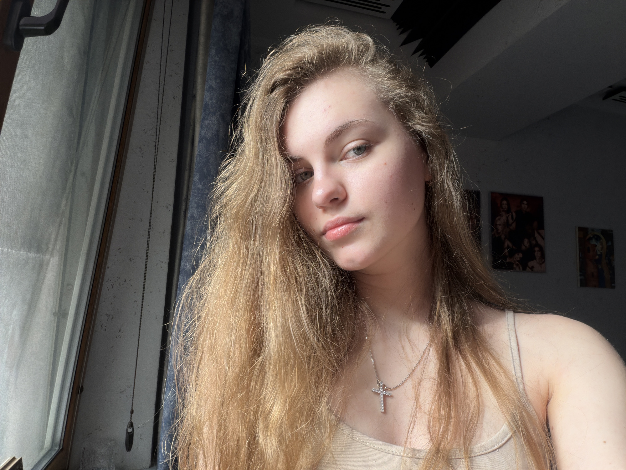 This is a portrait photograph of a young woman with long, blonde hair and fair skin. She is wearing a light-colored tank top and a silver cross necklace. The woman is looking towards the camera with a neutral expression. The background is dimly lit and appears to be an indoor setting, possibly a bedroom or living space. There are some framed pictures on the wall in the background. To the left of the woman, a window with a translucent curtain is visible, with the harsh light coming through it suggesting it is daytime. The scene does not provide specific details to identify the location in the city of Dnipro, Ukraine.