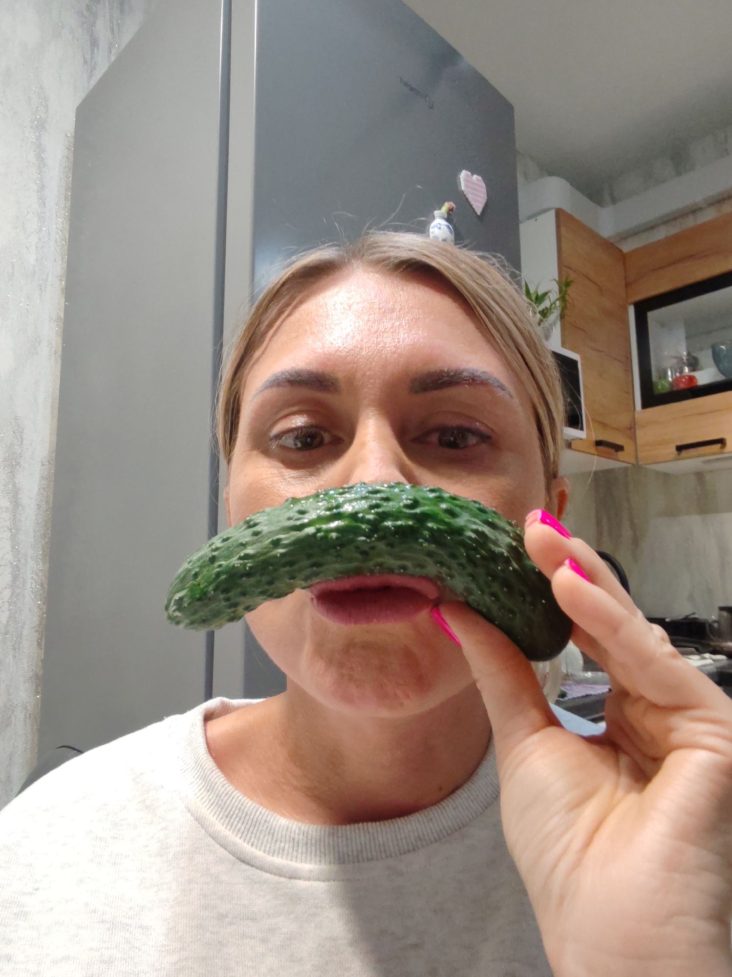 A close-up shot captures a woman holding a cucumber like a mustache in front of her face, indoors, possibly in a kitchen in Komsomolsk-on-Amur, Russia. The woman, with blonde hair pulled back, wears a light gray sweatshirt. Her right hand, adorned with bright pink nail polish, supports the cucumber. The background reveals a portion of a refrigerator, cabinetry with wooden accents, and a wall with textured wallpaper. The lighting suggests an indoor setting, possibly in the daytime.
