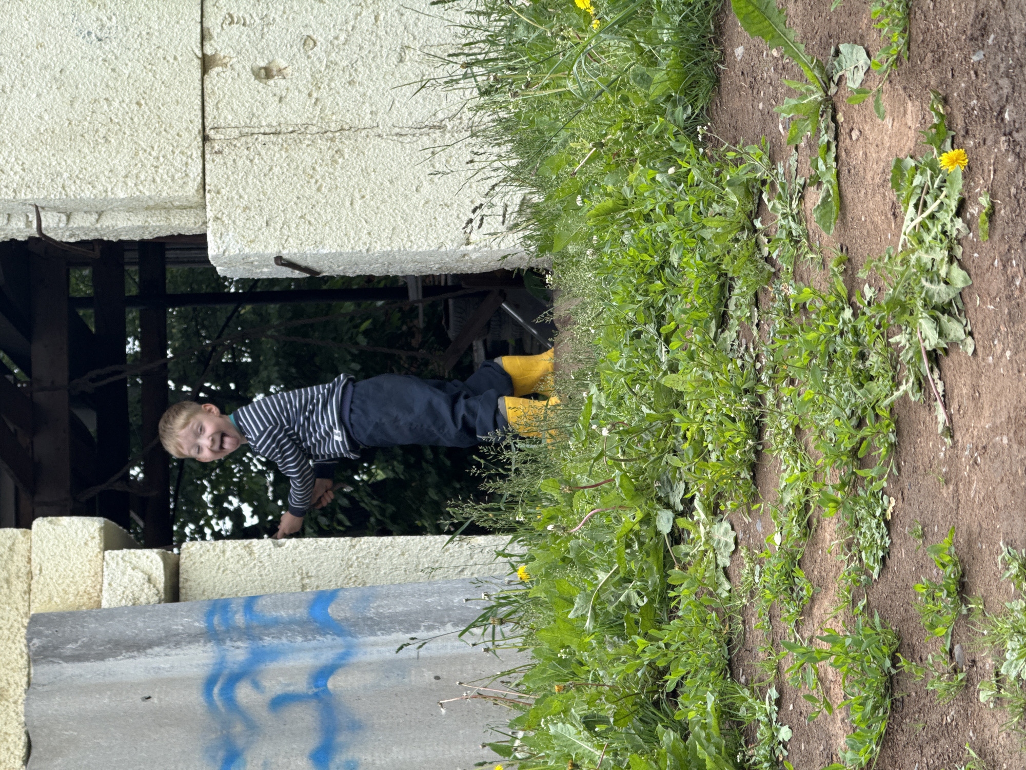 A young boy with blond hair sticks his tongue out at the camera while standing in front of a partially constructed building with light-colored foam blocks and a grey concrete panel. The boy is wearing a striped long-sleeved shirt, dark pants, and bright yellow rubber boots. He is positioned between two upright concrete or foam structures, with dense green vegetation and dirt in the foreground. The background shows trees and the interior of the structure, suggesting an outdoor, possibly rural, setting. The scene is likely in Nizhnyaya Chermoda, Russia.
