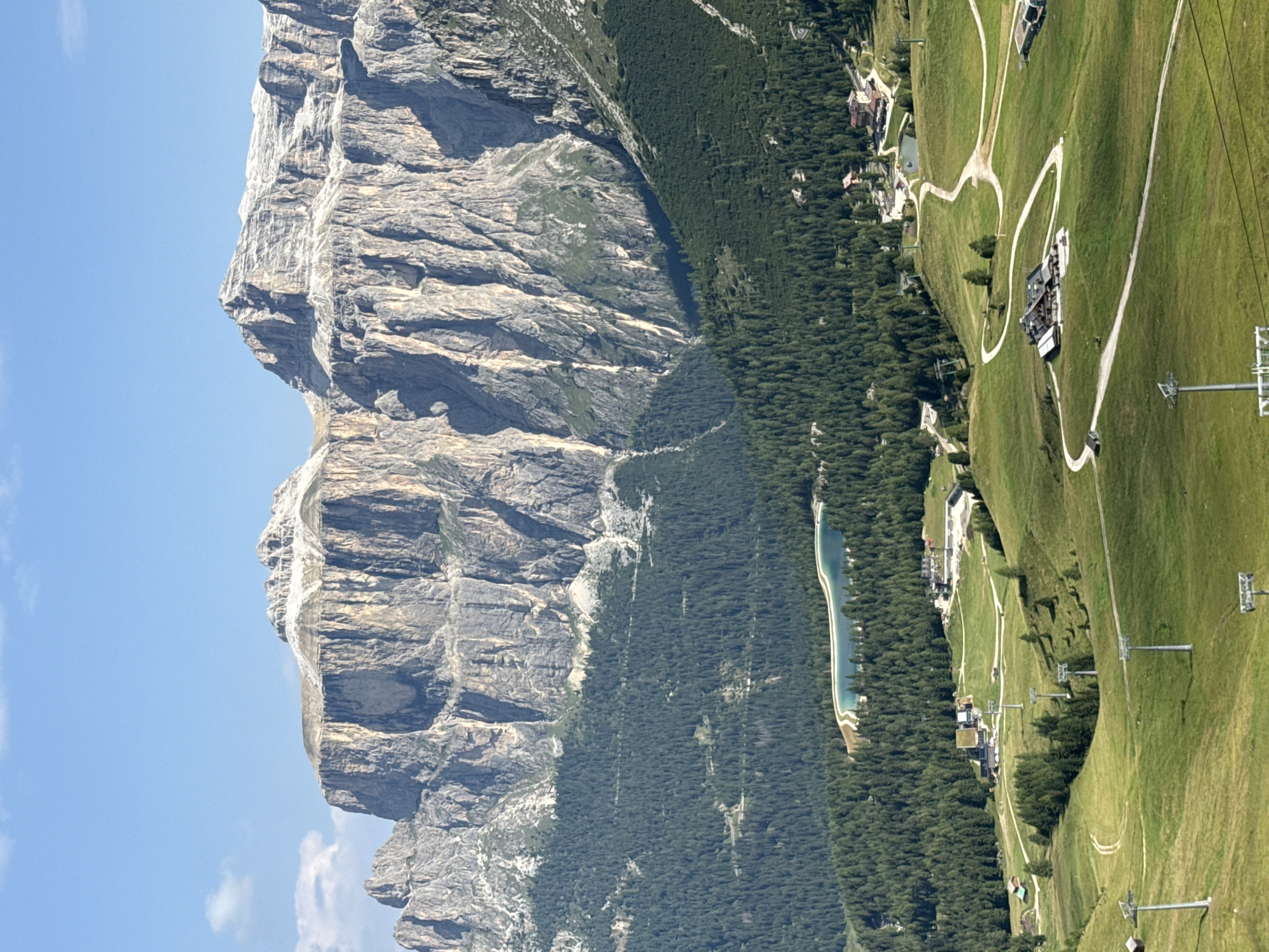 The image captures a majestic mountain landscape, likely in the Dolomites near Canazei, Italy. In the foreground, rolling green hills are crisscrossed by winding paths and dotted with sparse trees. Several ski lift towers are visible, suggesting a popular tourist destination, possibly for hiking in the summer and skiing in the winter. A few small buildings, possibly chalets or mountain huts, are nestled into the landscape. Further back, a dense, dark green forest blankets the lower slopes of towering, rugged mountains. These mountains are characterized by sheer rock faces, deeply fissured and displaying a palette of grays and browns. The peaks are capped with white, indicating snow or lighter colored rock. A small, serene lake with a distinctive aquamarine hue is visible nestled within the treeline, adding a touch of color to the scene. The sky above is a clear, bright blue with a few wispy clouds.
