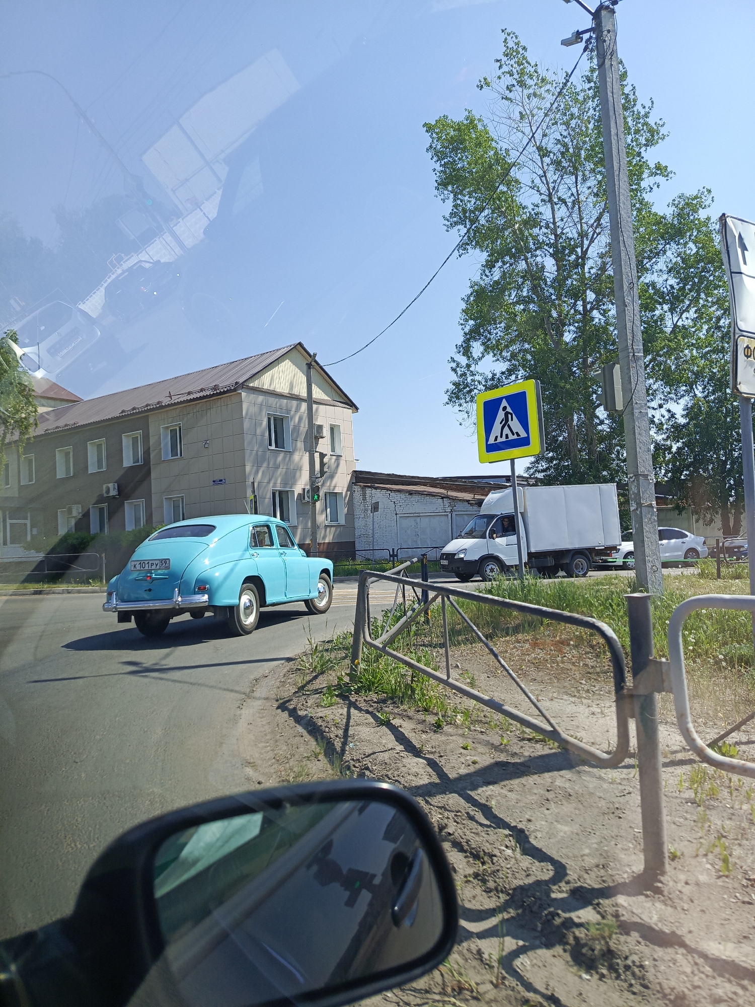 Captured from inside a vehicle, the image presents a street scene in Chaikovsky, Russia. A turquoise vintage car is seen driving on the street to the left. In the background, there is a multi-story building and other parked cars. A pedestrian crossing sign is visible on the side of the road, and a fence. The overall environment suggests a typical urban street corner on a sunny day. The presence of the car's side mirror on the bottom and a partial view of the car's interior frame the image and indicate it was taken from within a vehicle.