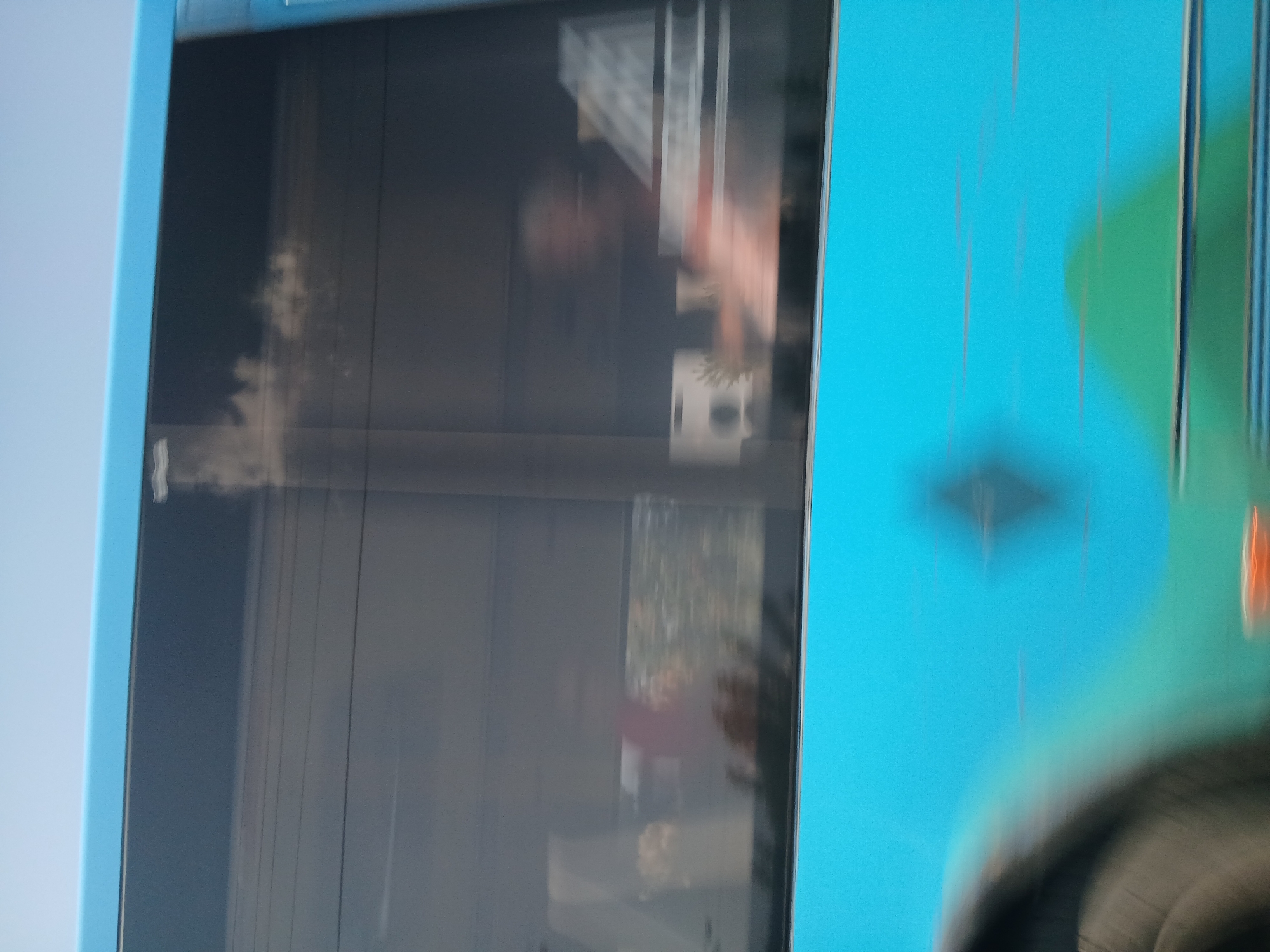 The image shows the side of a blue bus with tinted windows, captured in motion, as indicated by the blur. The sky is visible above the bus, and a reflection of clouds can be seen in the windows. Inside the bus, indistinct figures are visible through the windows, suggesting passengers. One figure appears to be a person with their arm raised. The bus itself has a blue and green design on its lower exterior. The overall scene is blurred, consistent with a photograph taken from a moving vehicle. The location is not identifiable from the image.