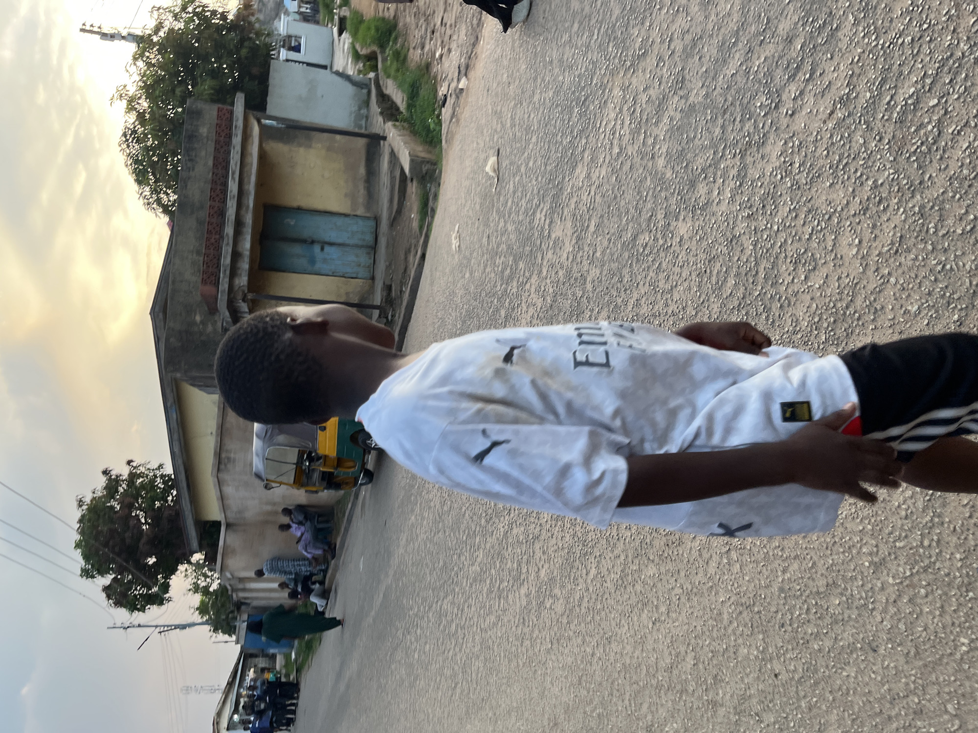 This image depicts an outdoor street scene in the city of Jos, Nigeria, at either dawn or dusk, characterized by the warm, diffuse light in the sky. In the foreground, a young, dark-skinned boy is partially visible from the left side, walking towards the right. He is wearing a white short-sleeved Puma t-shirt with "Emirates Fly..." text on the front, and black shorts with white vertical stripes. The road surface is unpaved or poorly paved, consisting of small, light-colored gravel.
In the midground, on the left side of the road, a group of people are gathered; some are seated on a low structure, while others stand. One individual, dressed in a long dark green garment, walks away from this group towards the left. A yellow and green motorized tricycle, commonly known as a Keke Napep, is parked on the right side of the road, positioned behind the walking boy. On the right, a single-story building with light-colored, weathered walls, a flat roof, and a distinctive blue double-door entrance is prominent. A decorative pattern of red bricks adorns the top edge of this building. Overhead power lines traverse the sky, and sparse trees and vegetation are visible in the background, including a large tree situated behind the building on the right. The sky exhibits soft cloud formations with a warm, yellowish hue near the horizon, gradually transitioning to a lighter blue overhead.