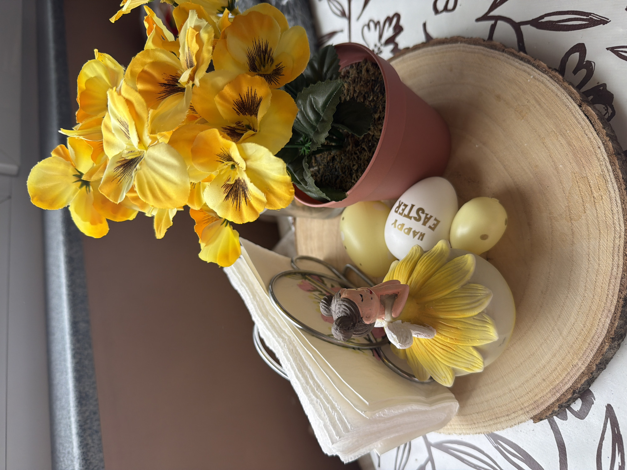A top-down, tilted view captures a still life arrangement. A pot of yellow and brown pansies sits on a circular wooden slab with bark edging. Next to the pot are Easter eggs; one white egg has "HAPPY EASTER" printed on it in gold. A napkin holder containing white napkins is topped with a small figurine and a yellow flower. The wooden slab rests on a surface with a white and brown floral pattern. The left side of the image reveals a brown surface adjacent to a gray surface. The setting is likely a household interior. There's no indication of being in Buhovo, Bulgaria.