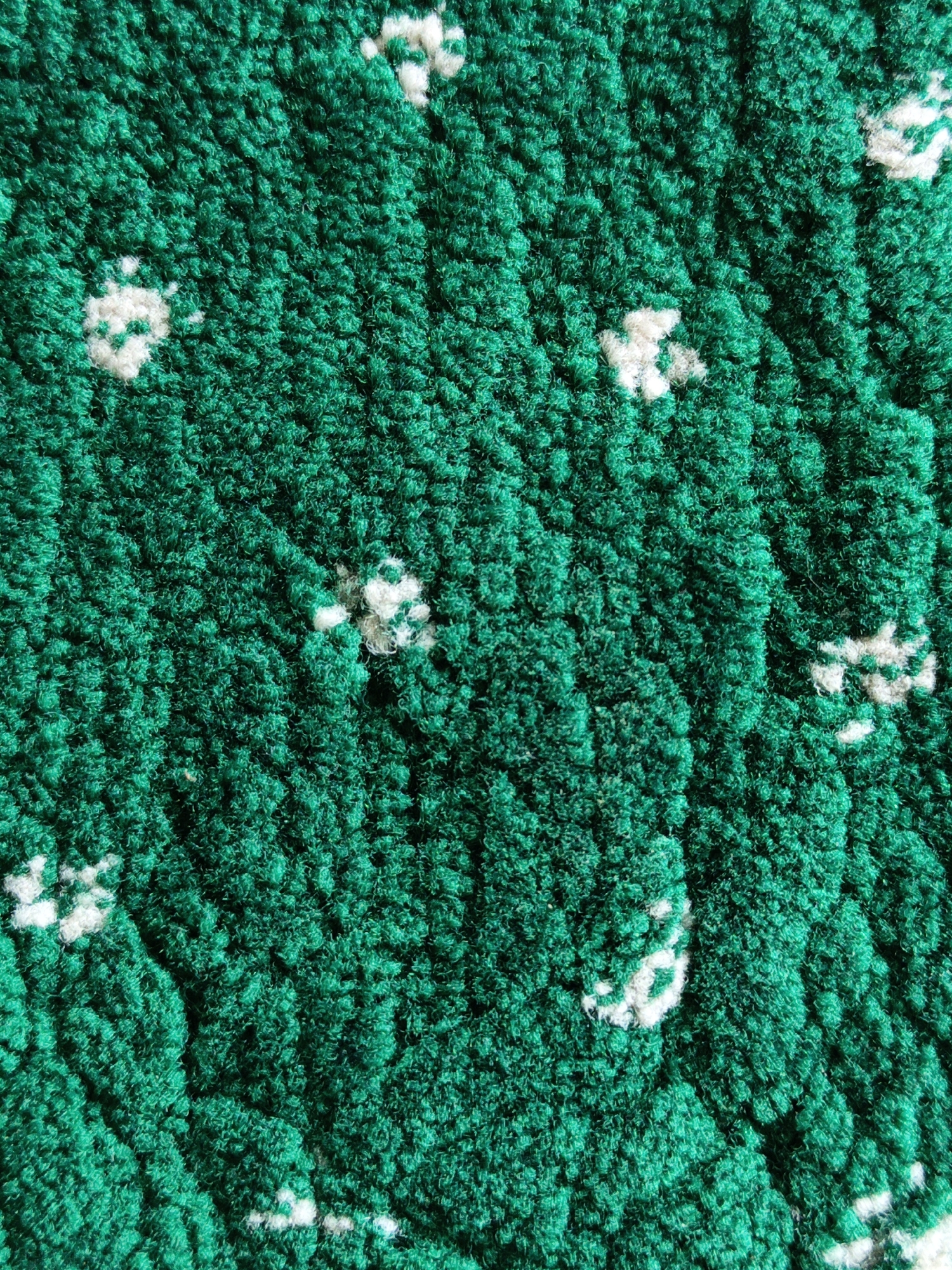 A close-up, high-angle, full-frame shot showcases the texture of a green shag rug with scattered white floral or clover-like patterns. The rug's fibers are dense and plush, creating a soft, inviting surface with shadows and highlights that emphasize its depth. The white patterns are irregular in shape and placement, appearing as if they were embroidered or woven into the green fabric. No people or actions are visible in the image, and the context of Kotabumi, Indonesia, is not apparent from the visual information provided. The image is purely focused on the textile's surface.