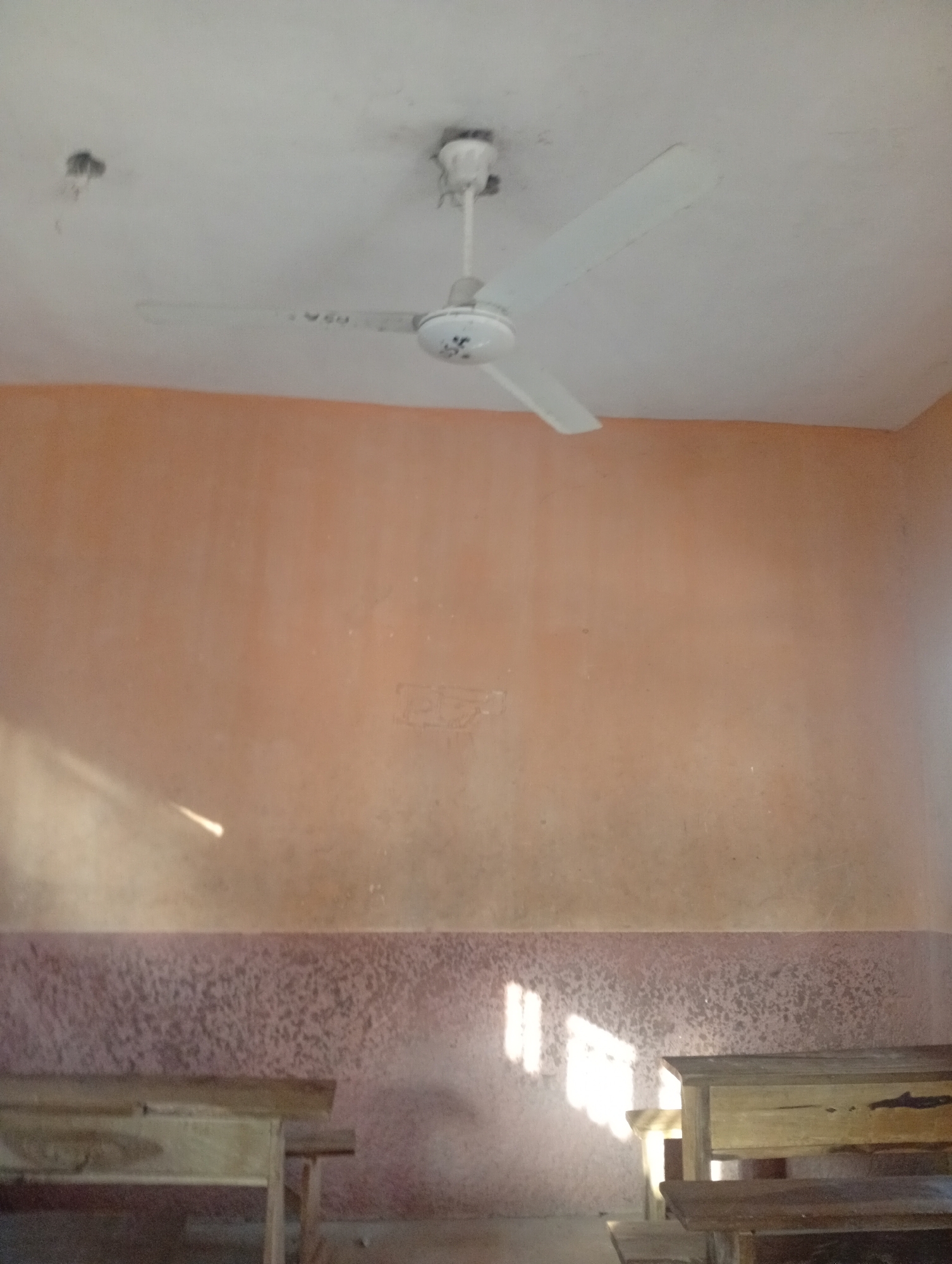 The media file depicts the interior of what appears to be a classroom. A white ceiling fan with three blades is prominently featured, hanging from the ceiling. The walls are painted in a peachy-orange hue, with a textured, darker band along the lower portion of the walls. Sunlight streams in from the left, casting light patterns on the wall and the lower textured band. Two wooden desks are partially visible in the foreground, suggesting a learning environment. The overall impression is of a simple, perhaps aged, room. The lighting suggests it is daytime, with the sun's angle indicating it could be morning or afternoon. The setting appears to be Jalingo, Nigeria, as indicated by the location context. There is no visible text within the image.