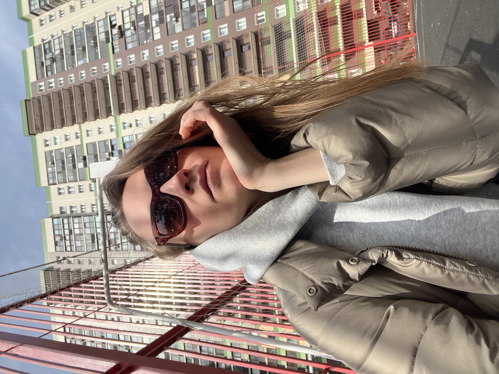 A young woman wearing sunglasses, a grey hoodie, and a beige puffer jacket poses for a selfie. She is standing on a bridge or elevated platform with a red metal railing. In the background, there is a tall apartment building with light green, beige, and brown exterior walls, featuring multiple windows and balconies. The location is Khimki, Russia, indicated by the architectural style of the building. The weather appears to be sunny and clear.