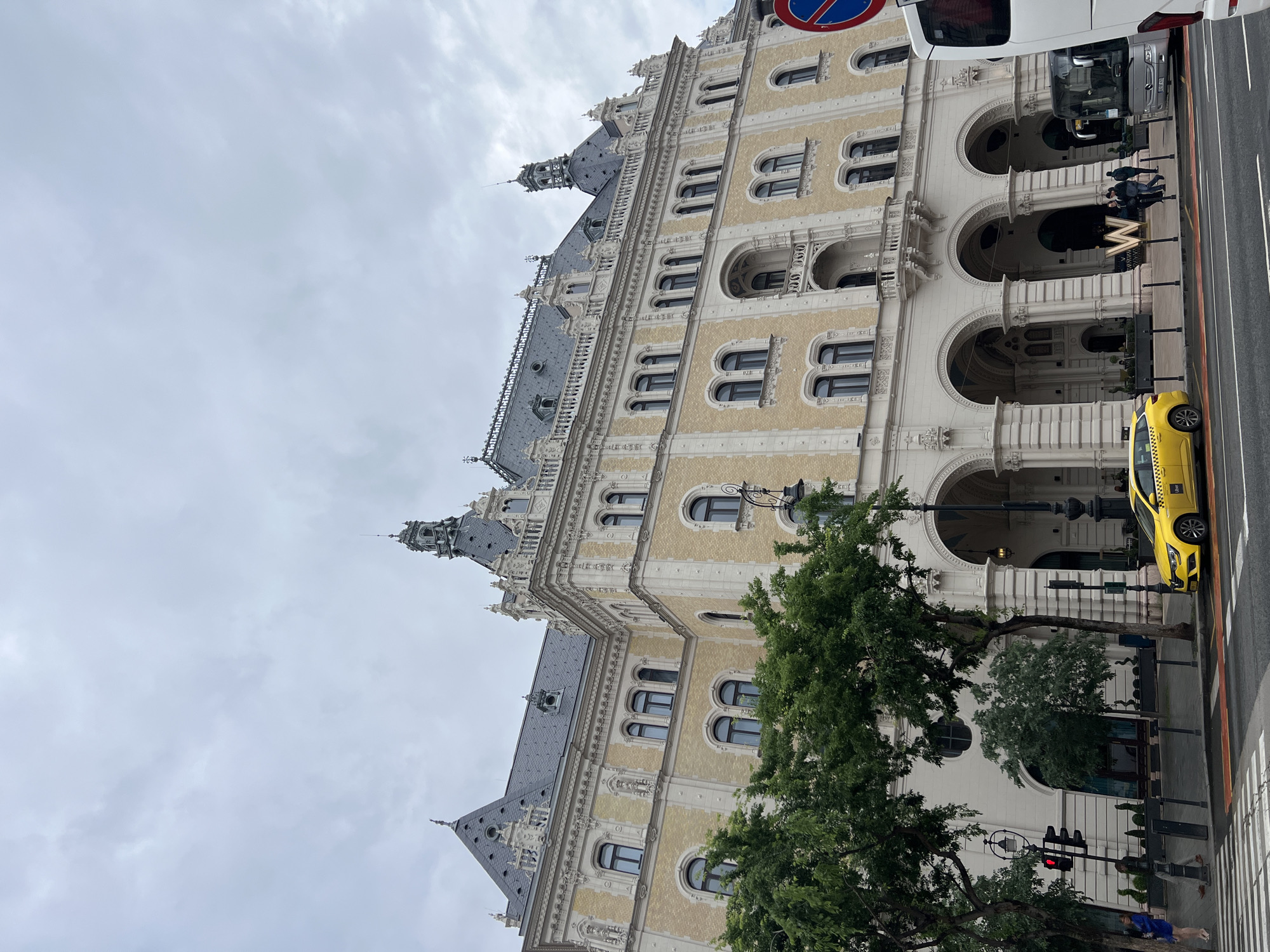 The image features a view of the Gresham Palace in Budapest, Hungary. The building is prominently displayed, showcasing its ornate architecture with multiple floors, arched windows, and intricate detailing. The sky is overcast, providing a muted light across the scene.
In the foreground, trees partially obscure the base of the building. A yellow taxi is parked along the street. Pedestrians can be seen walking along the sidewalk. A white van is parked parallel to the taxi. A road sign is visible in the upper right corner of the image. The "W" logo for the W Hotel is visible on the building. A traffic light is showing red, indicating the traffic status.