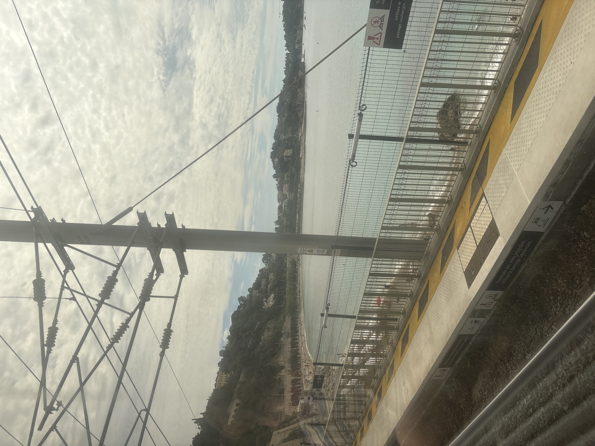 The image shows a view from a train moving along the coast of Villefranche-sur-Mer, France. The railway line runs parallel to the shoreline, offering a glimpse of the beach and the Mediterranean Sea. The foreground features train infrastructure, including overhead power lines and a platform edge with safety barriers and warning signs in French regarding train crossings. The background includes the cityscape of Villefranche-sur-Mer, extending up the hillsides, and the cloudy sky above.