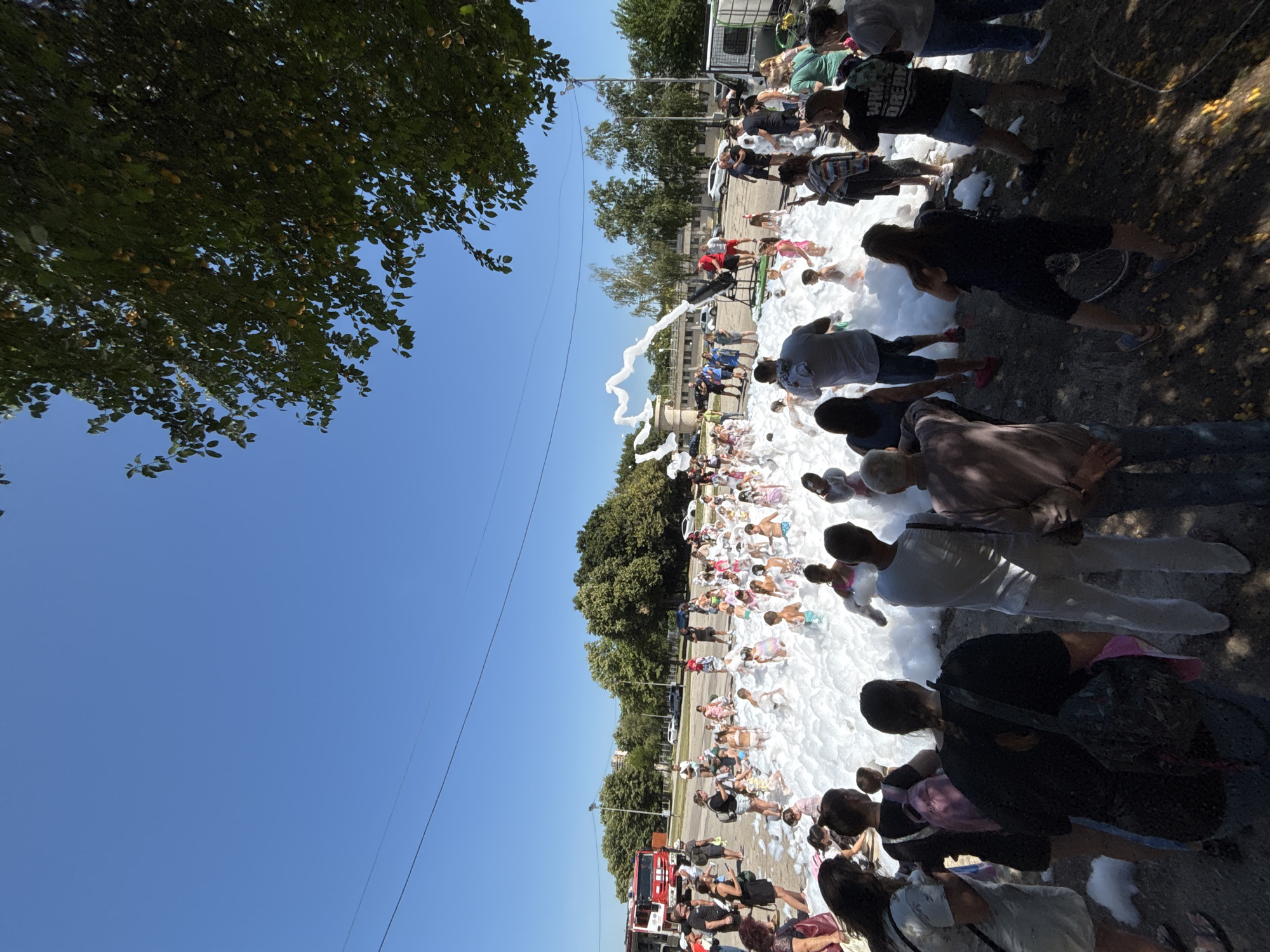 The image depicts a foam party with numerous children playing in a large pile of foam. Several adults are observing the children from the perimeter. In the background, a red fire truck is visible, suggesting a possible connection to emergency services or a community event. The scene takes place outdoors under a clear blue sky. Trees and buildings are present in the background, indicating an urban or suburban environment. The location is the city of Vratsa, Bulgaria.