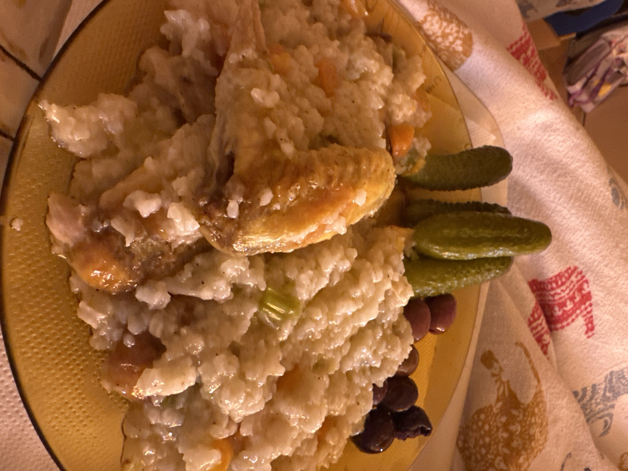 A yellow plate of food sits on a patterned tablecloth. The plate contains a mound of rice, with diced vegetables mixed in. There are two pieces of cooked chicken on top of the rice. To the right of the rice and chicken are three small, green pickles and a cluster of dark olives. The tablecloth has a white background with red, blue, and yellow patterns of horses and other figures. The scene appears to be indoors, possibly a home or restaurant. The location is not discernable based on the image alone.