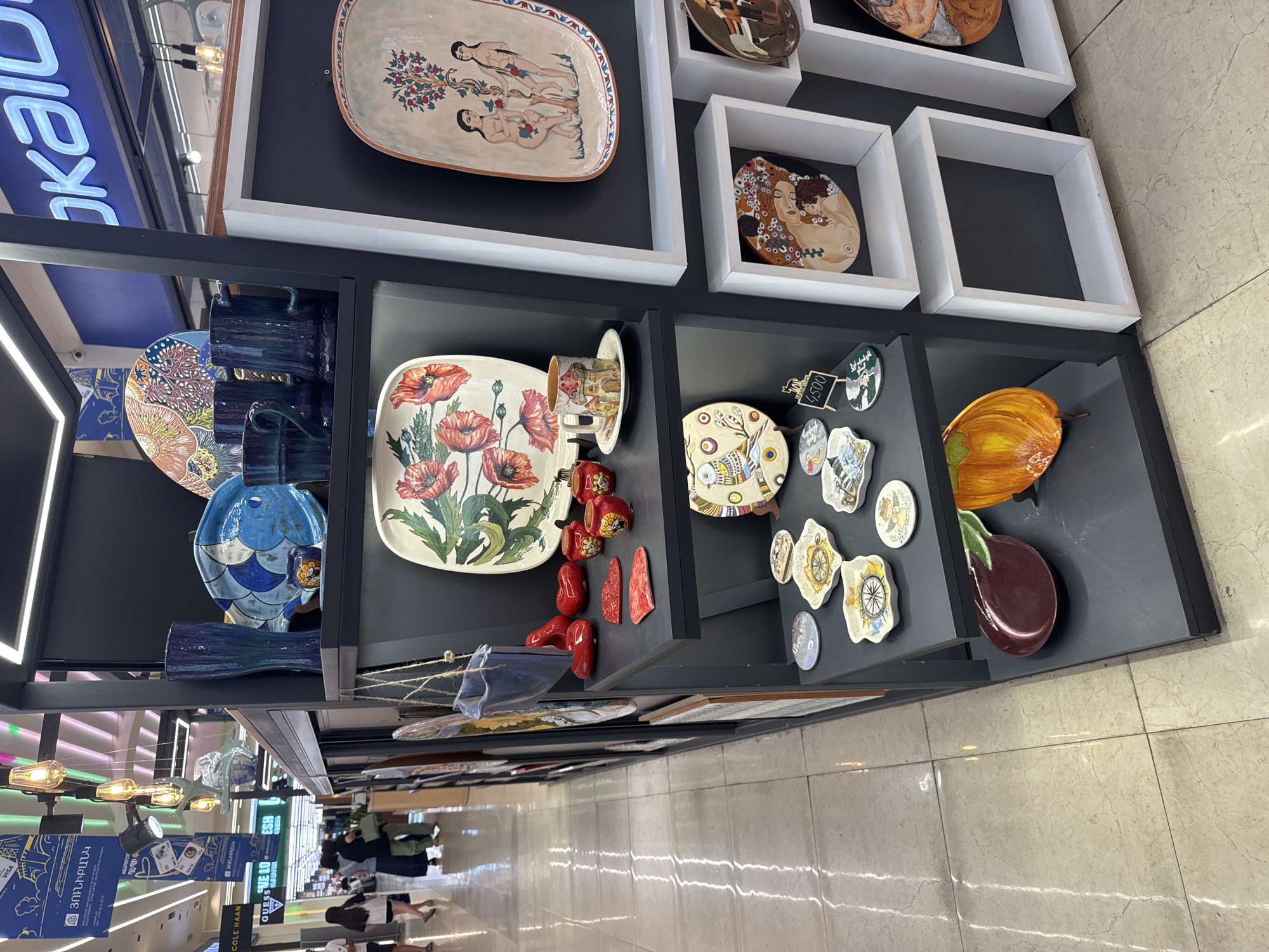 The image depicts a display of decorative ceramic items, possibly in a shop or market stall, located in Yerevan, Armenia. The stall features multiple shelves filled with various ceramic pieces, including plates, cups, and figurines. Prominently displayed are several large plates with painted floral and figurative designs, as well as smaller decorative dishes and mugs. The items are arranged on dark gray shelves, with some items hung from the ceiling. In the background, blurred figures of shoppers and storefronts are visible, suggesting a retail environment. The lighting appears to be a mix of overhead fluorescent lights and decorative pendant lights. The overall impression is of a vibrant display of artisanal ceramics.