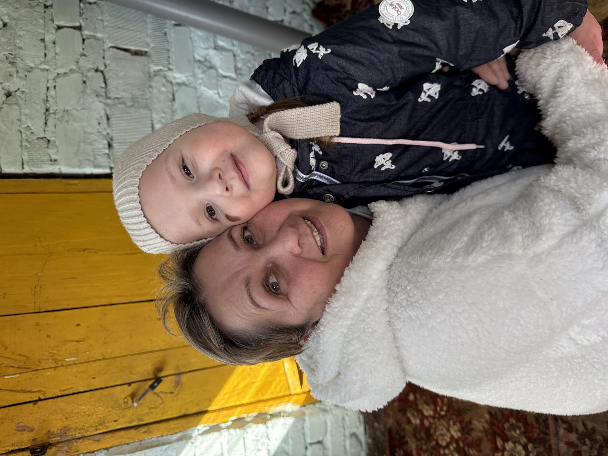 A young child and an adult, presumably the child's grandmother, are embracing in an outdoor setting in Kipchak, Russia. The child is wearing a winter hat and jacket with a distinctive pattern, while the adult is bundled in a white, fleece-like jacket. The background features a building with a yellow wooden section and a white brick portion. A metal pipe runs horizontally across the top of the frame. A section of patterned carpet is visible in the lower right corner of the image.