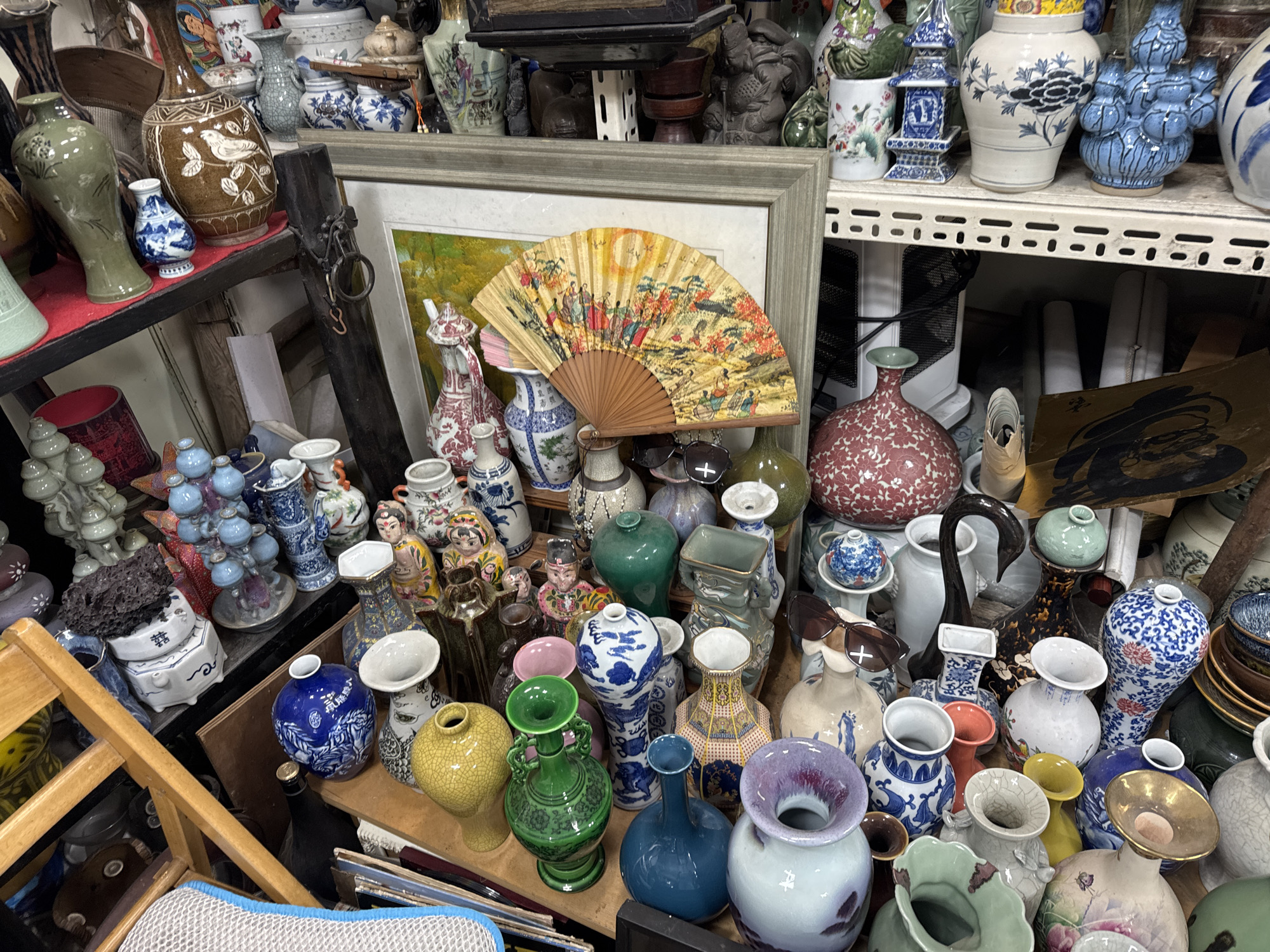 An eye-level, close-up shot shows a cluttered antique shop in Seoul, South Korea. Numerous vases of various sizes, colors, and designs occupy most of the frame. In the background, more shelves with vases are visible. A framed painting and a folding fan with Asian artwork rest among the vases. A wooden chair is partially visible in the lower left corner. Sunglasses sit on top of some vases, adding to the disorganized appearance.